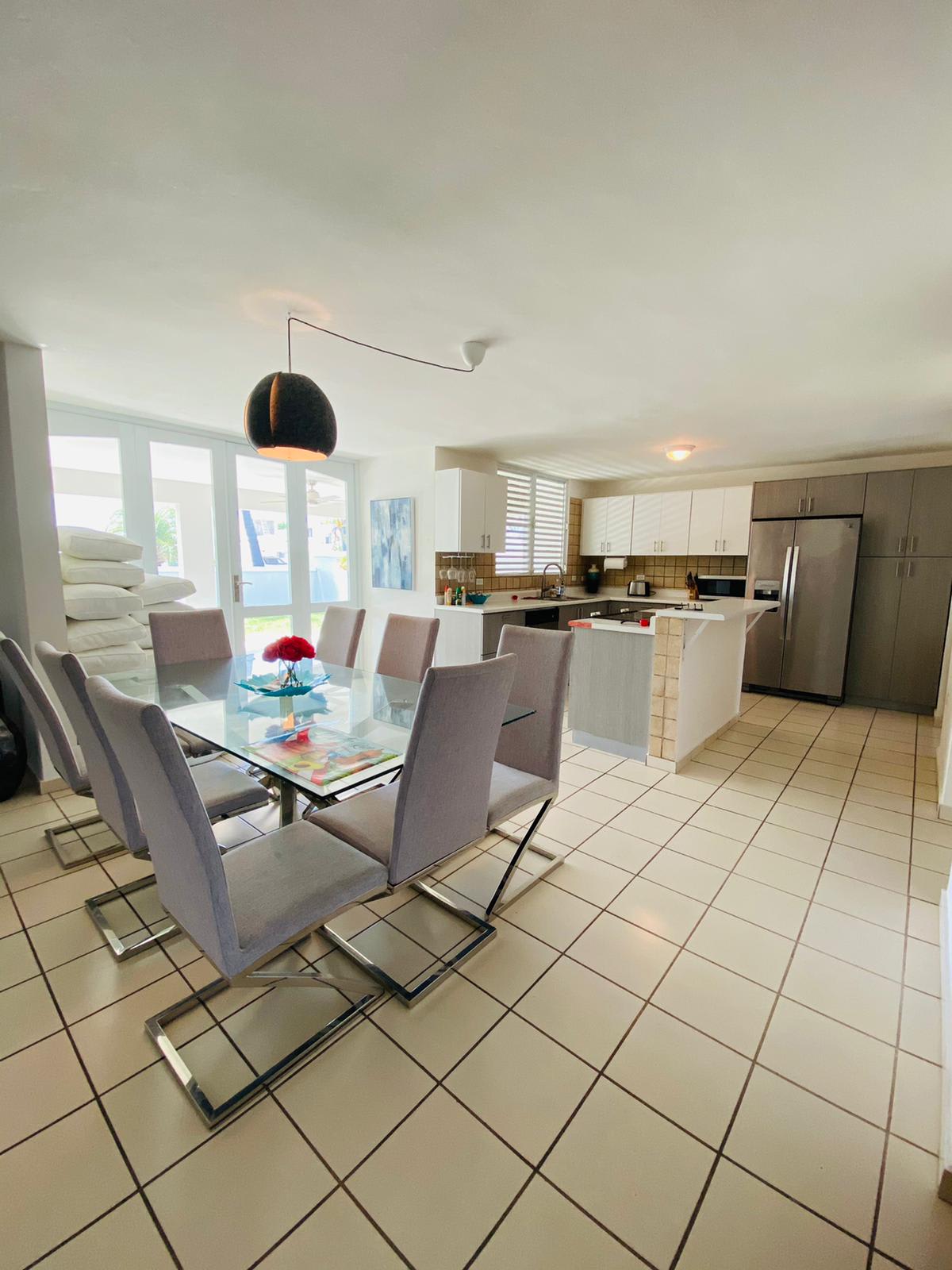 A bright and airy dining room with a glass table and chairs in front of a kitchen.