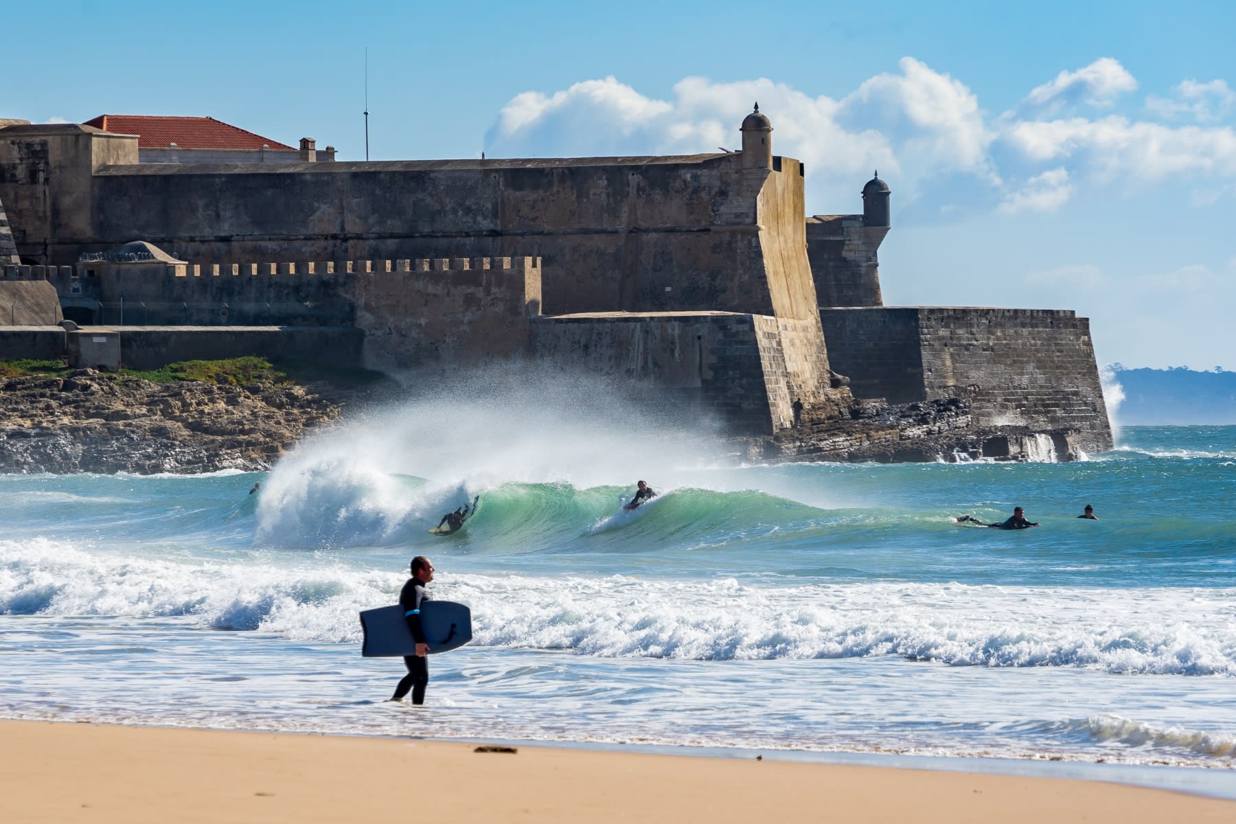 Surfers at a sandy beach below a stone seaside fort.