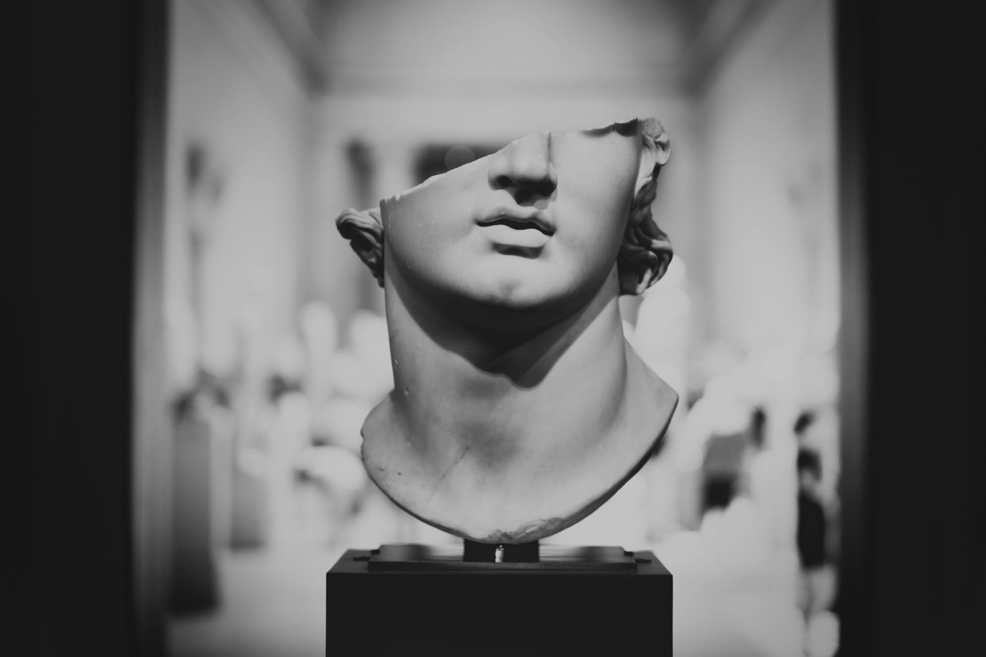 Classical sculpted bust on a pedestal, shown in black and white with a gallery of sculptures behind it.