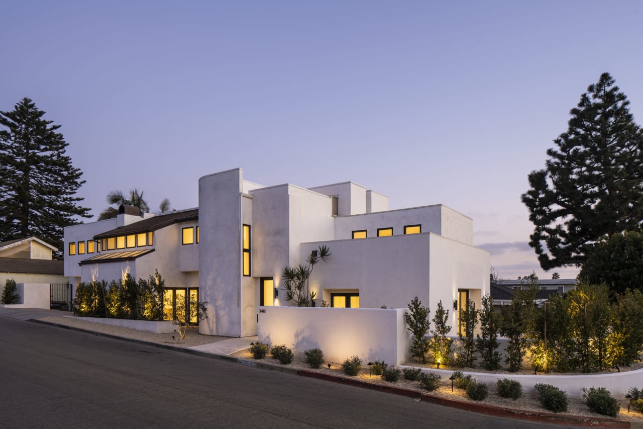 A modern white house with a minimalist design sits on a sloping street, illuminated by warm lights against a twilight sky.