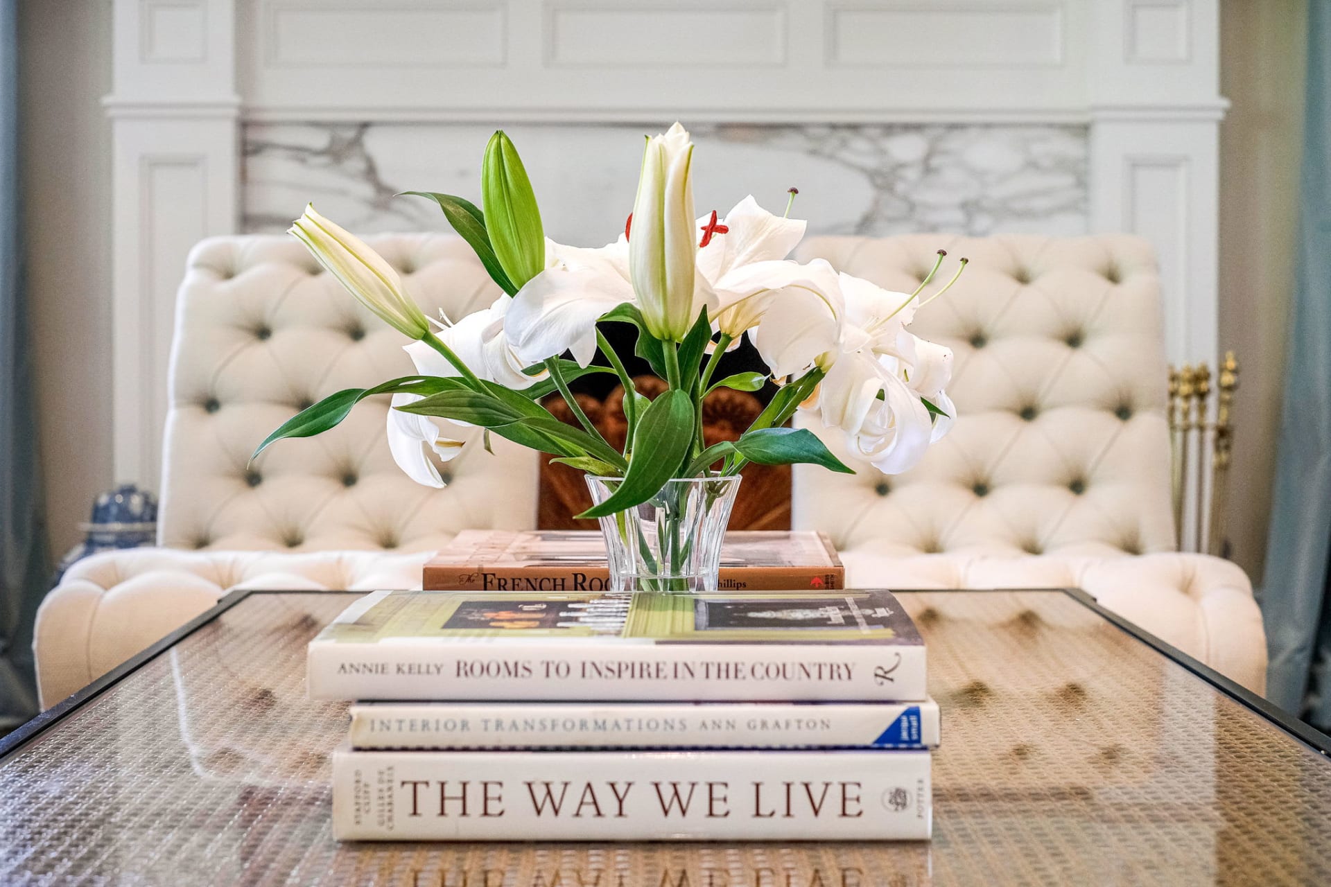  A beautifully styled glass coffee table in a living room features a clear vase filled with elegant white lilies and lush green foliage as its centerpiece. 