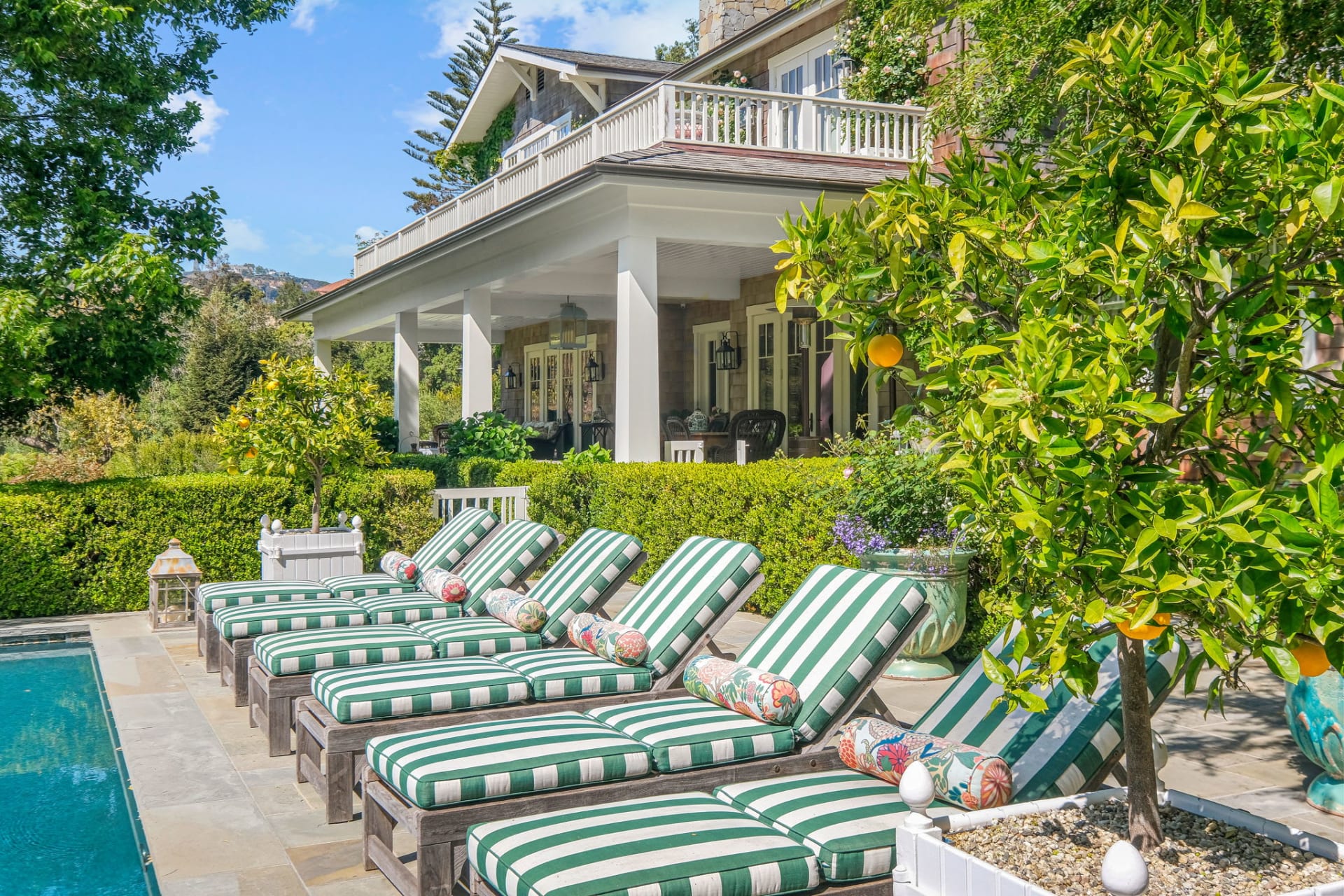  A luxurious outdoor scene features a row of six wooden lounge chairs with green and white striped cushions, positioned beside the sparkling blue water of a swimming pool. 