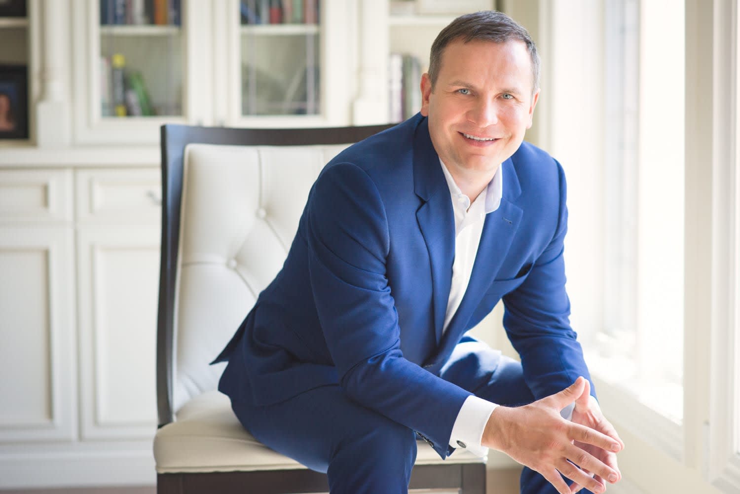 Man in a blue suit seated on a white chair in a bright interior.