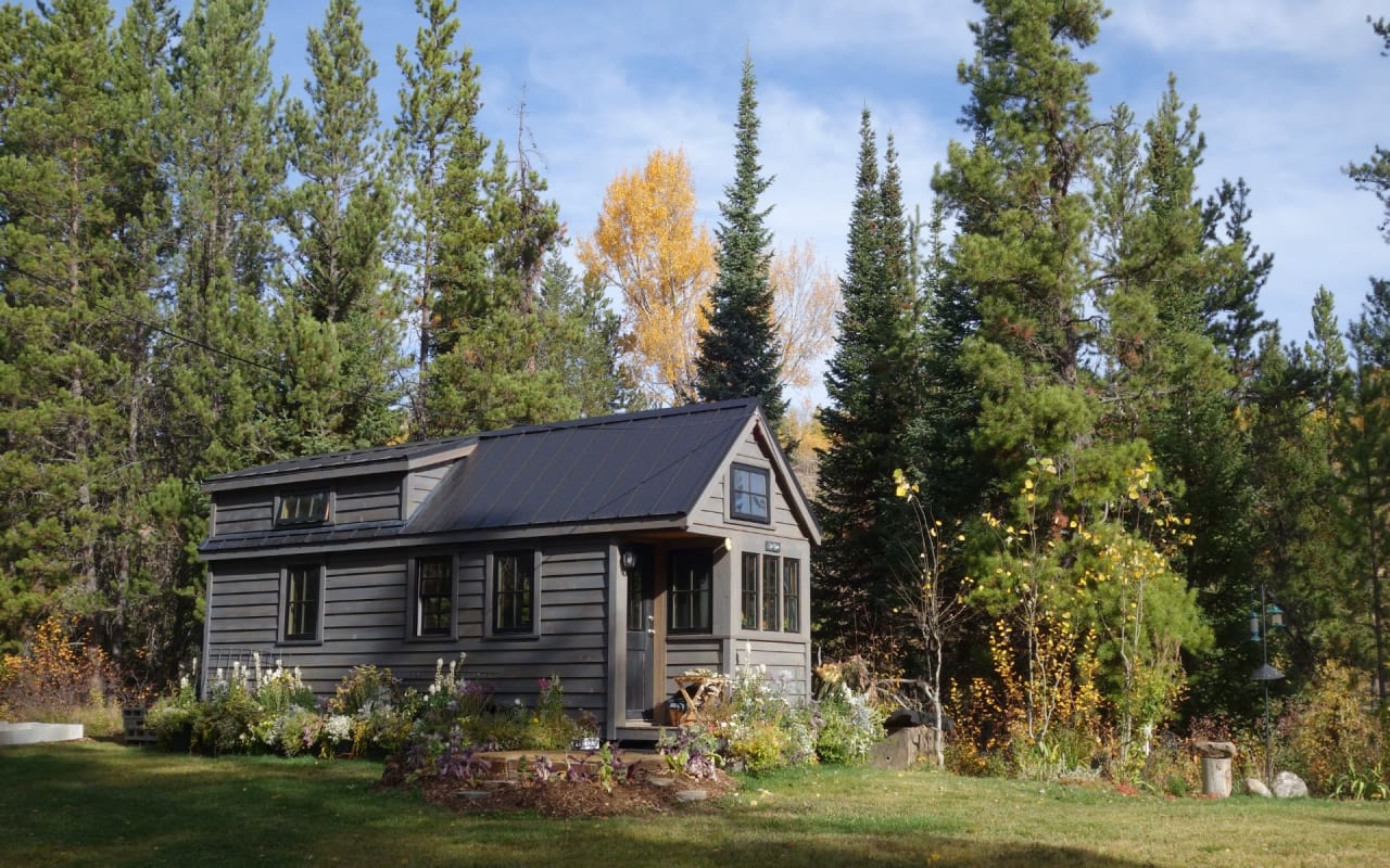 Wooden house with black metal roof, small porch, surrounded by flowers and trees in a forest.