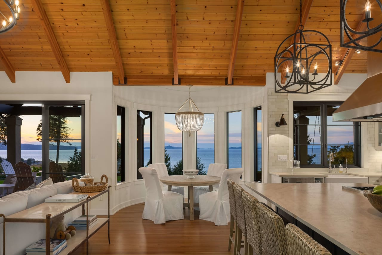 Cozy dining area with a chandelier, white chairs, and a wooden table, set against large windows with ocean views.