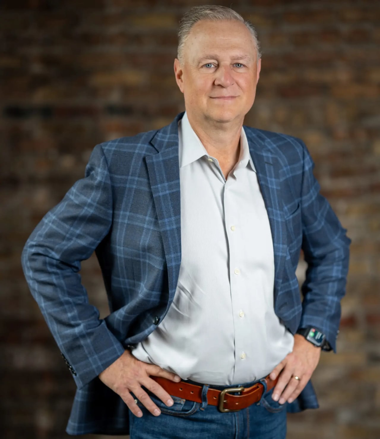 Lance Kammes in a blue checkered blazer with hands on hips standing against a brick wall.