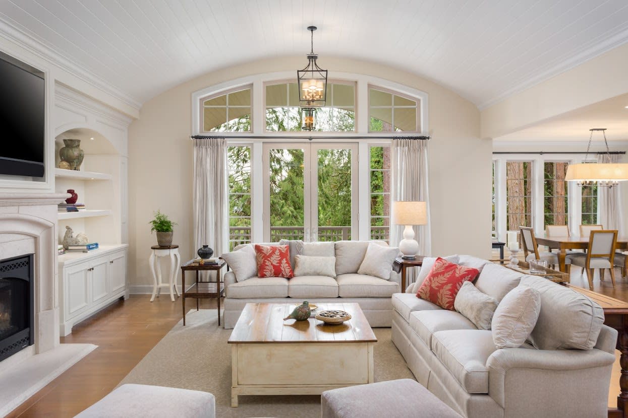 A living room with various furniture with cream-color décor, a stone fireplace, and large windows.