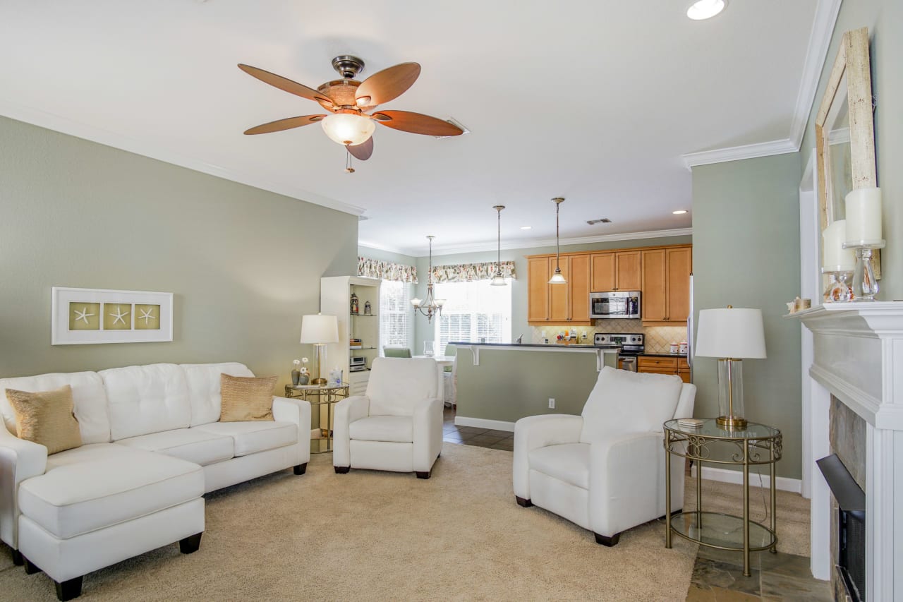 An interior view of a cozy living room with light-colored walls, carpeted floors, and a ceiling fan. The room is furnished with comfortable seating and a fireplace.