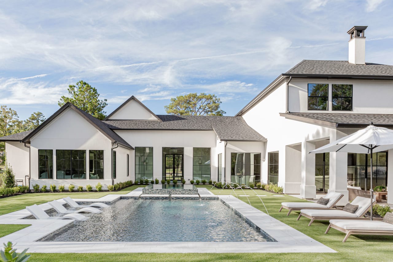 A luxurious white two-story house with a large swimming pool in front, surrounded by a patio with lounge chairs and tables