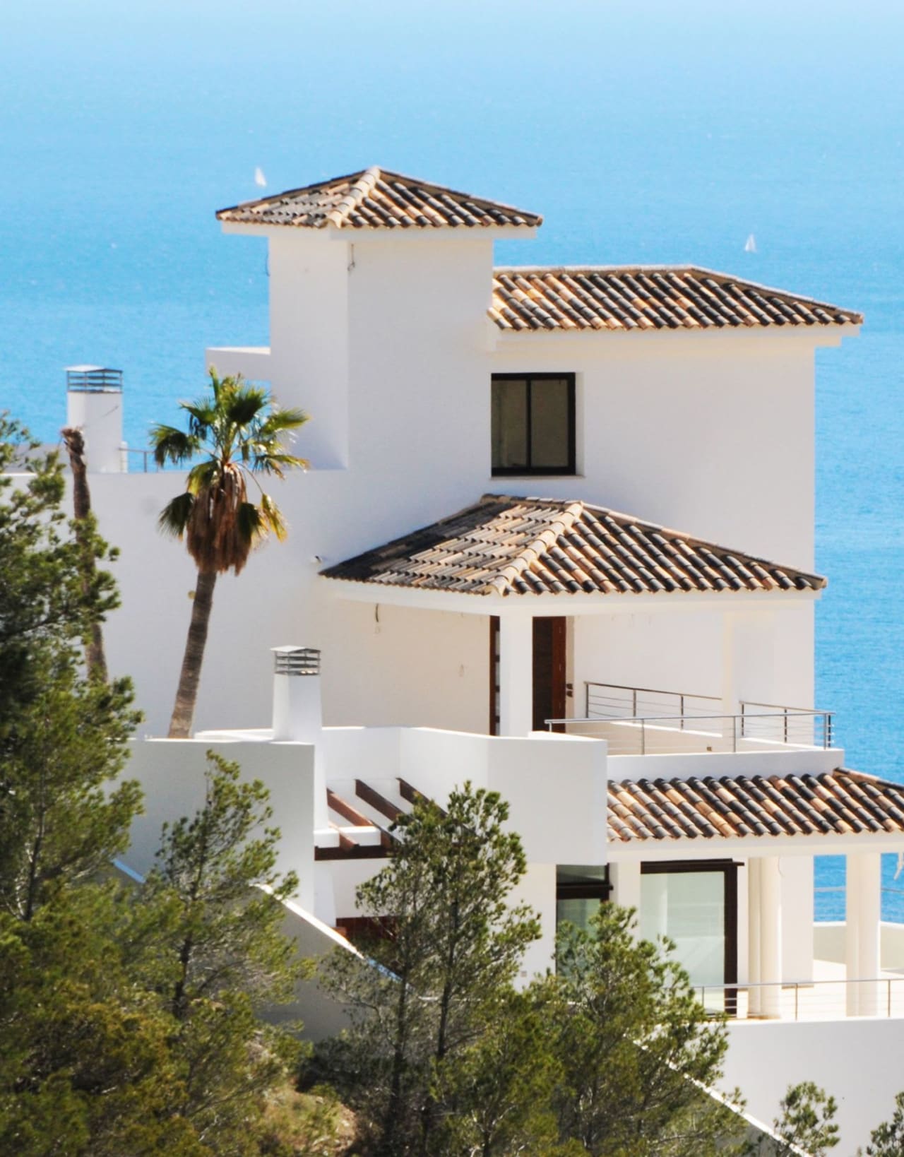 White house with red roof in Mediterranean region on a hill by the sea, with palm trees, balcony, and distant sailboats.