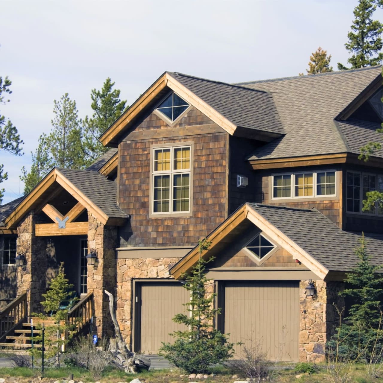 A large brown house with two garages in front, nestled in a wooded area.