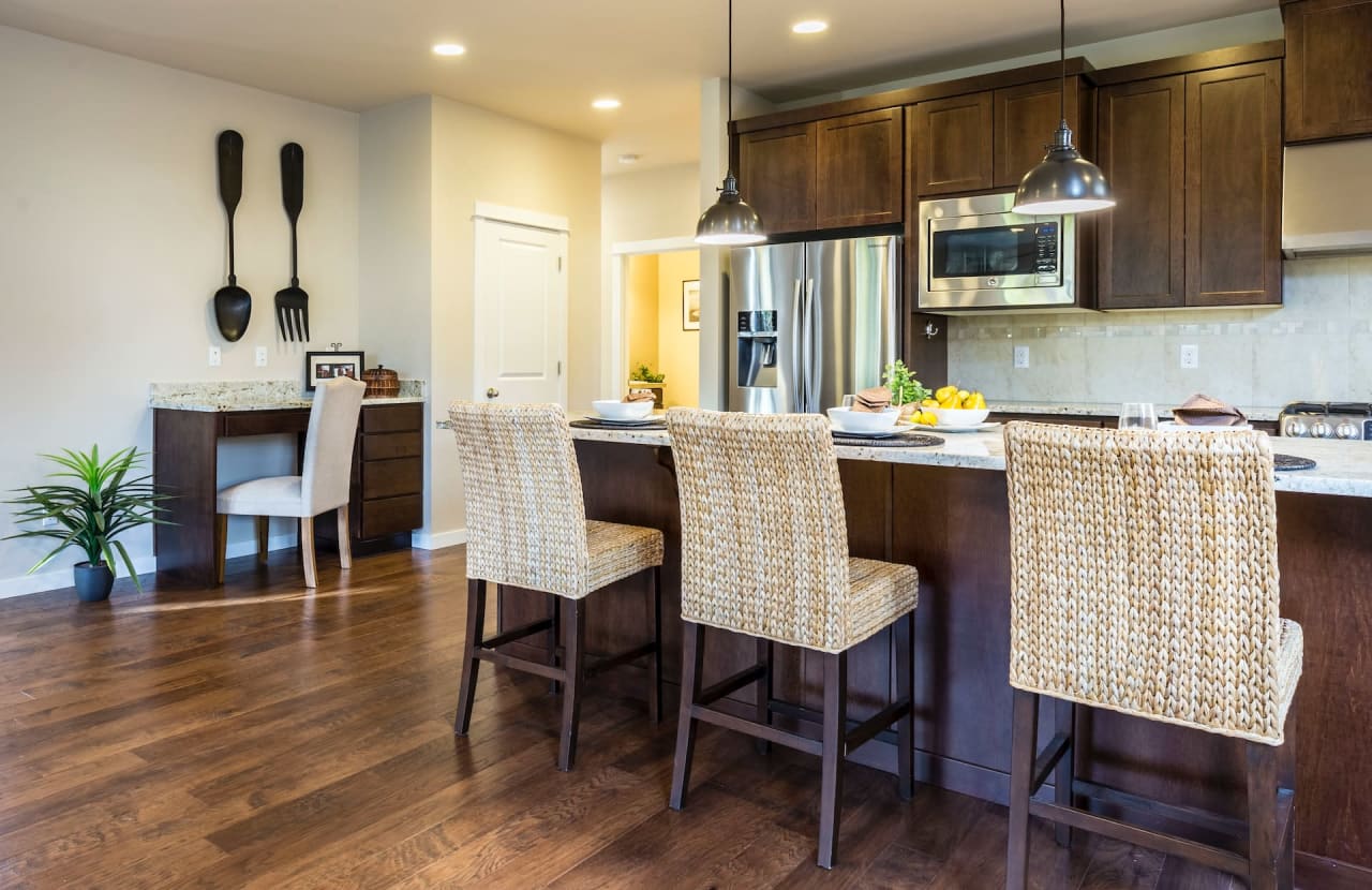 Cozy kitchen with dark wood cabinets, woven bar stools, stainless steel appliances, and oversized wall utensils as decor.