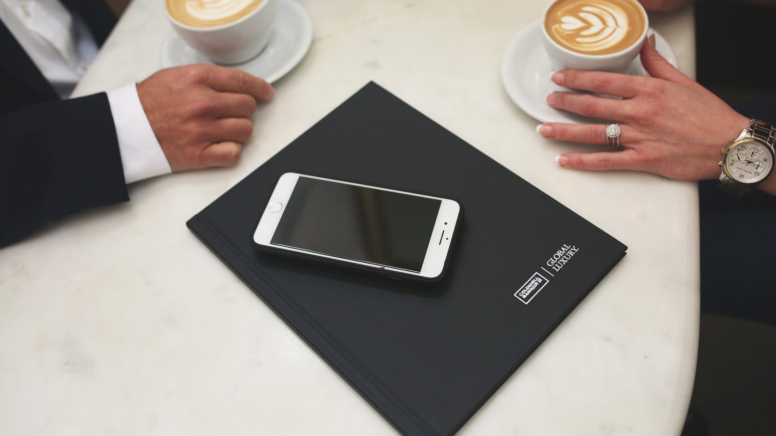 Two people with lattes at a white marble table, with a smartphone resting on a black Coldwell Banker Global Luxury presentation folder between them.