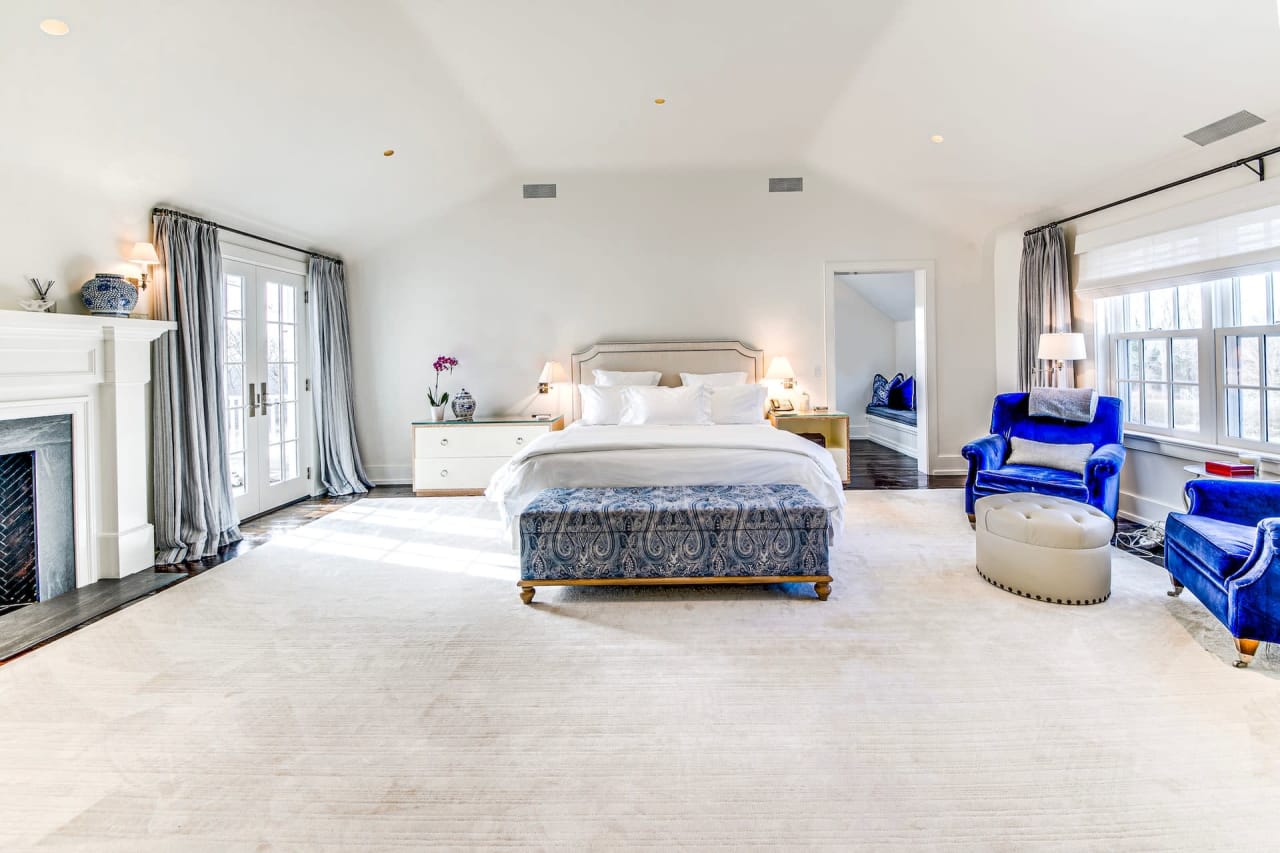 A cozy bedroom with a light-colored bed, wooden nightstand, fireplace, mirror, balcony door, and two windows.