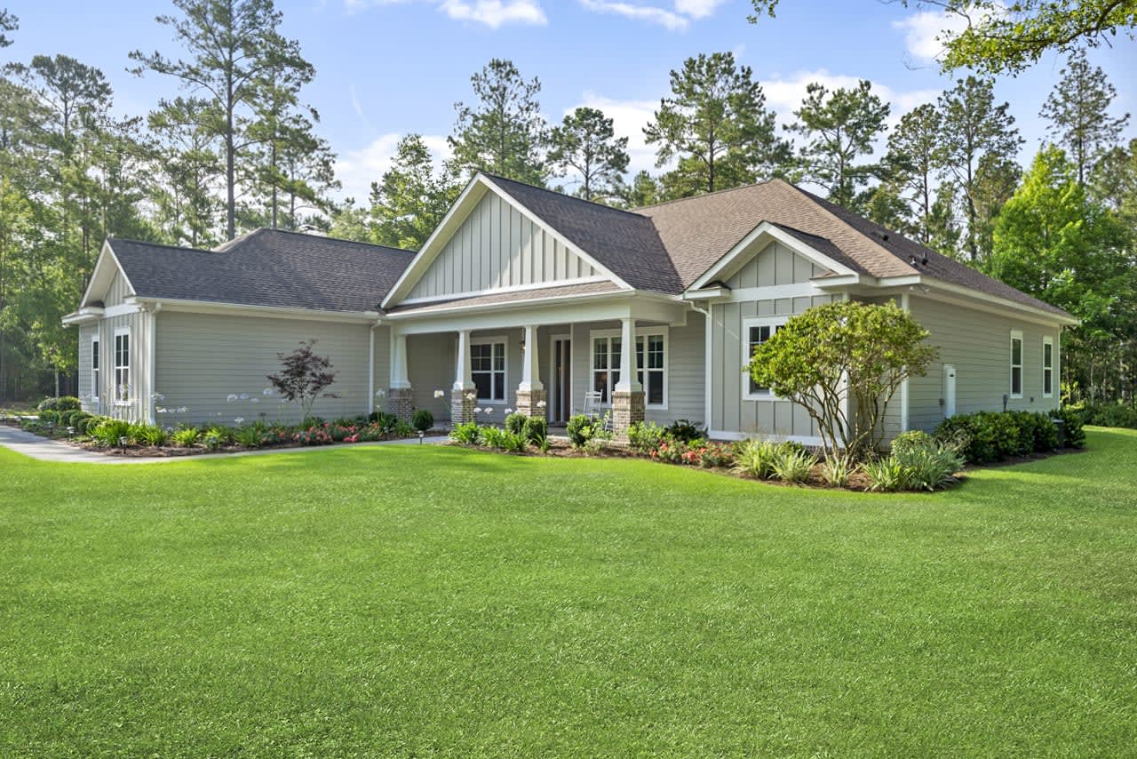 An exterior view of a large, single-story house with a well-manicured lawn, trees, and surrounding greenery. The house has a traditional architectural style.