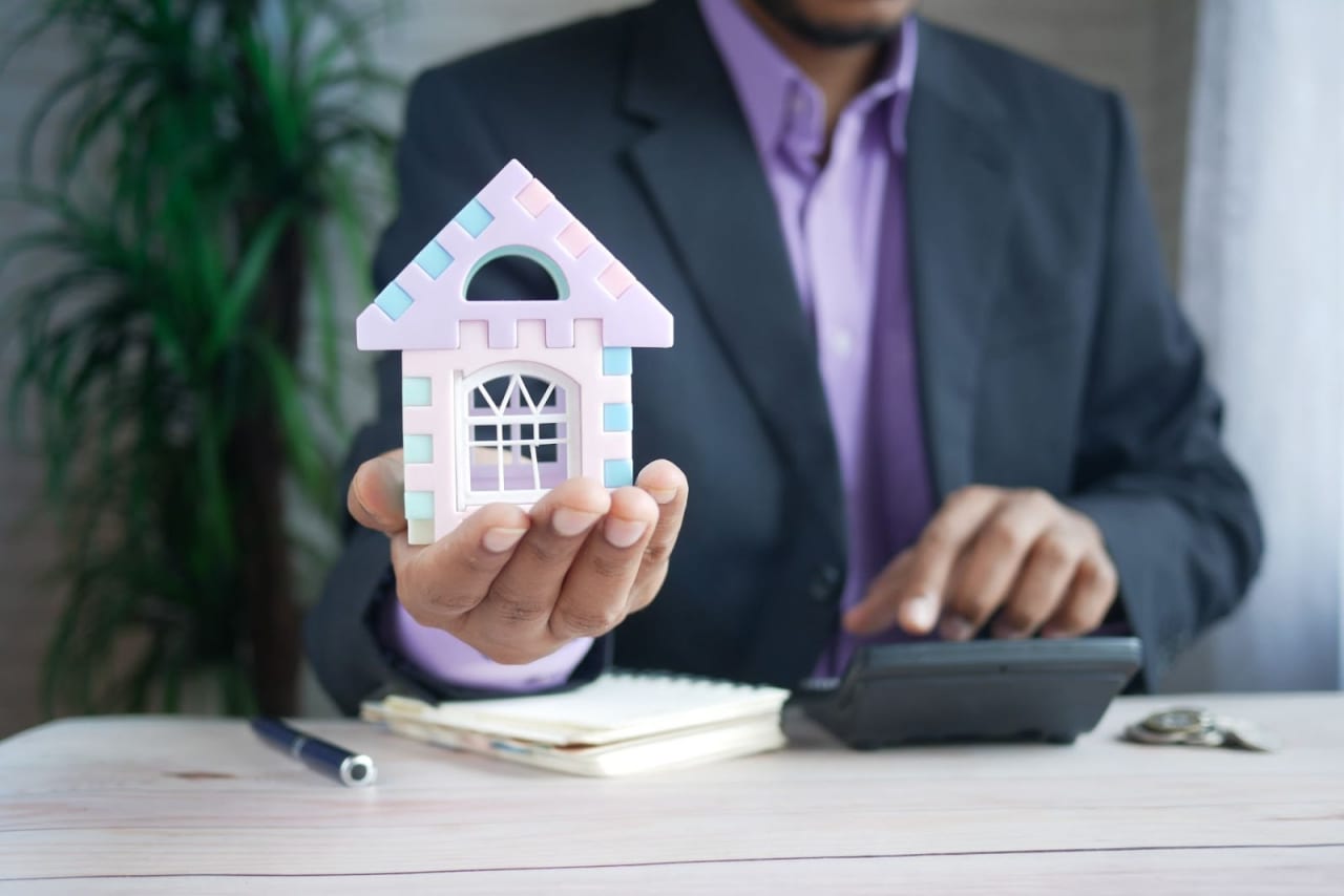 A Man Holding a Tiny House with a Notebook, Pen, and Calculator on the Table