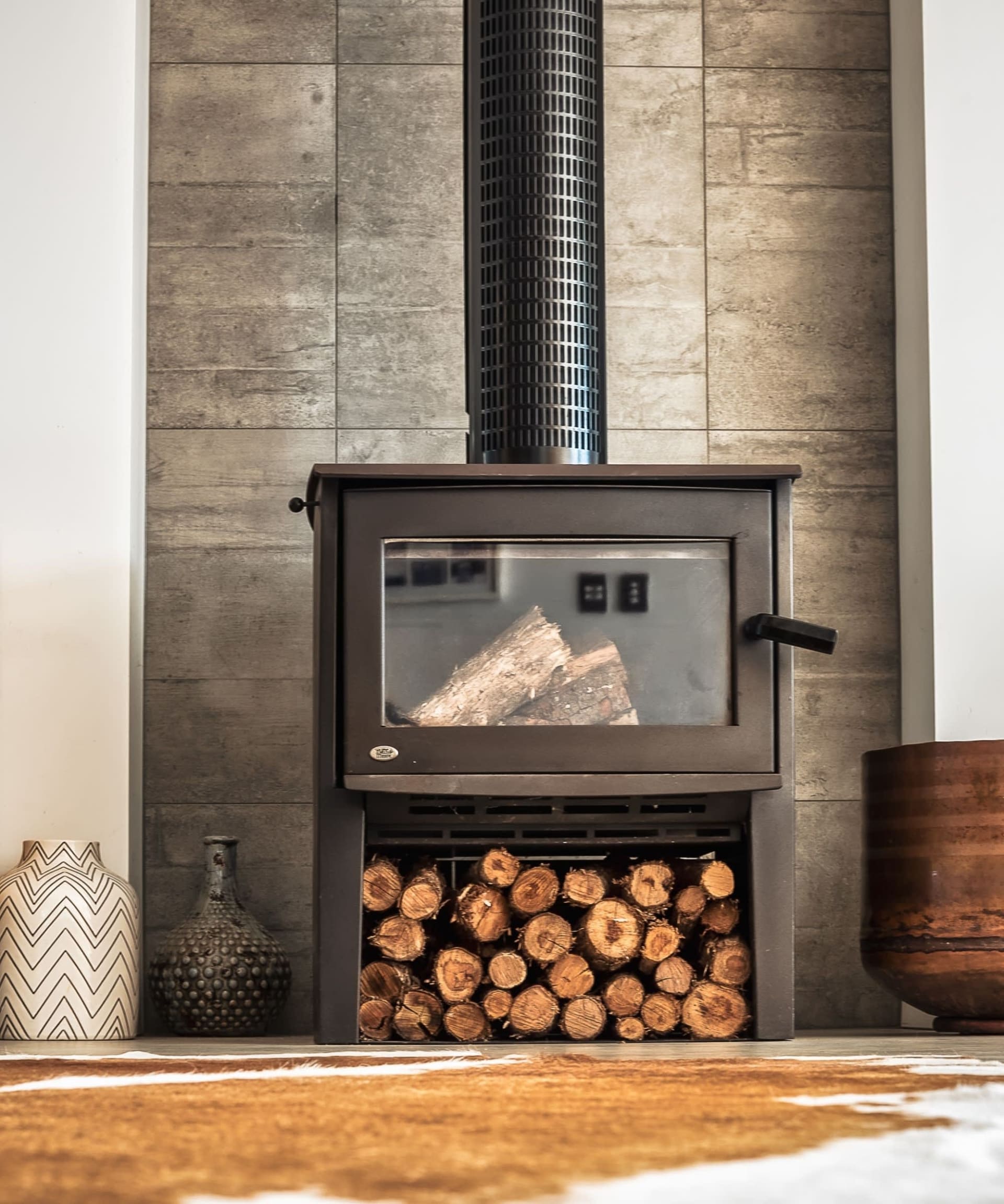 Black wood-burning stove against a stone-tiled wall, with firewood stacked beneath it.