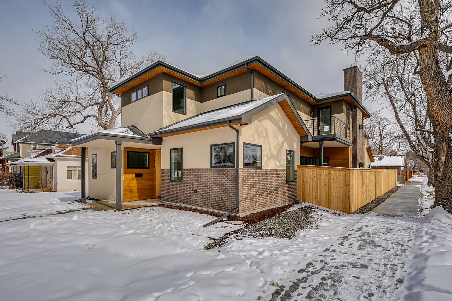  A contemporary two-story house with a mix of light tan stucco, dark brown siding, and light brick accents, standing in a snow-covered landscape.