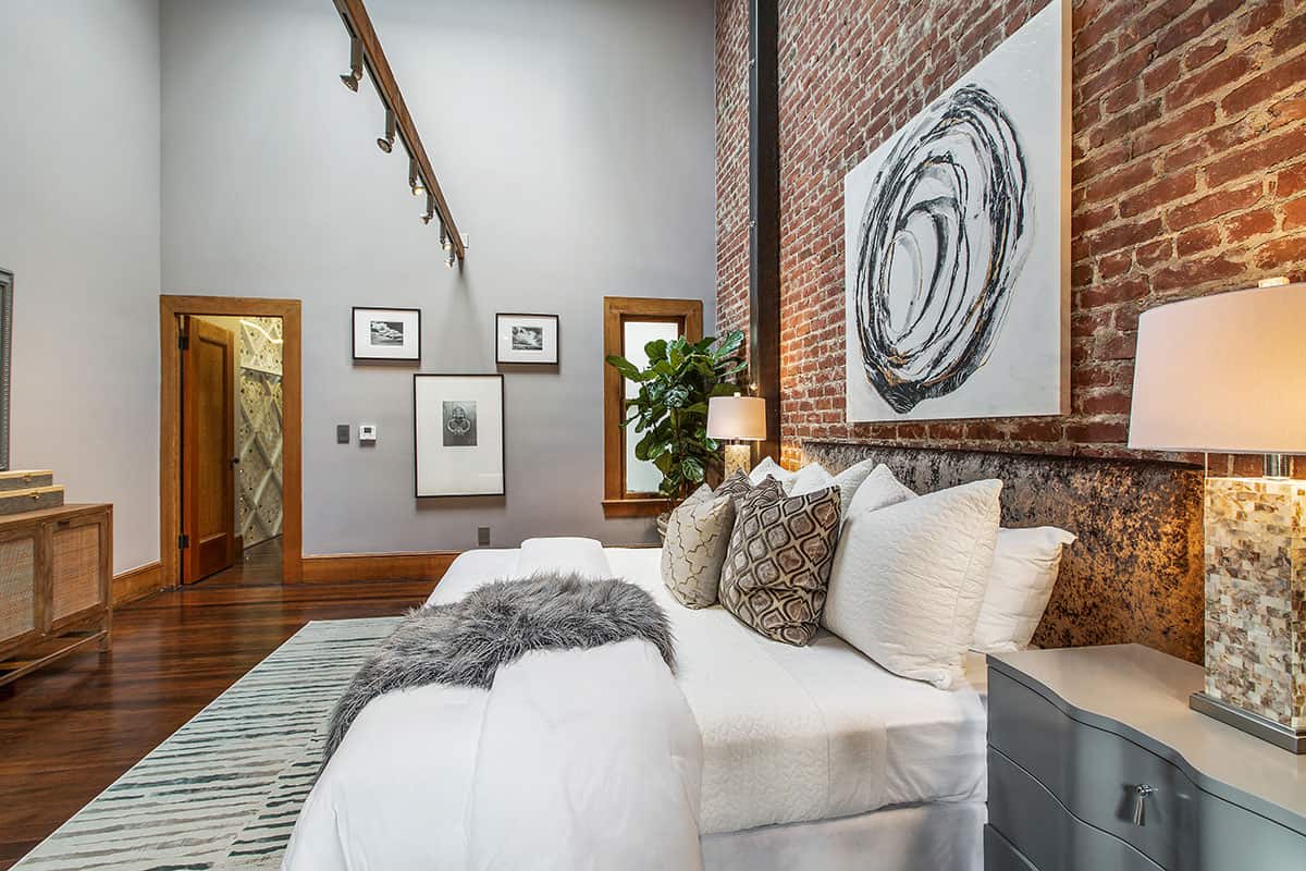 Bedroom with a white bed, exposed brick wall, dark wood floors, and oversized abstract artwork.