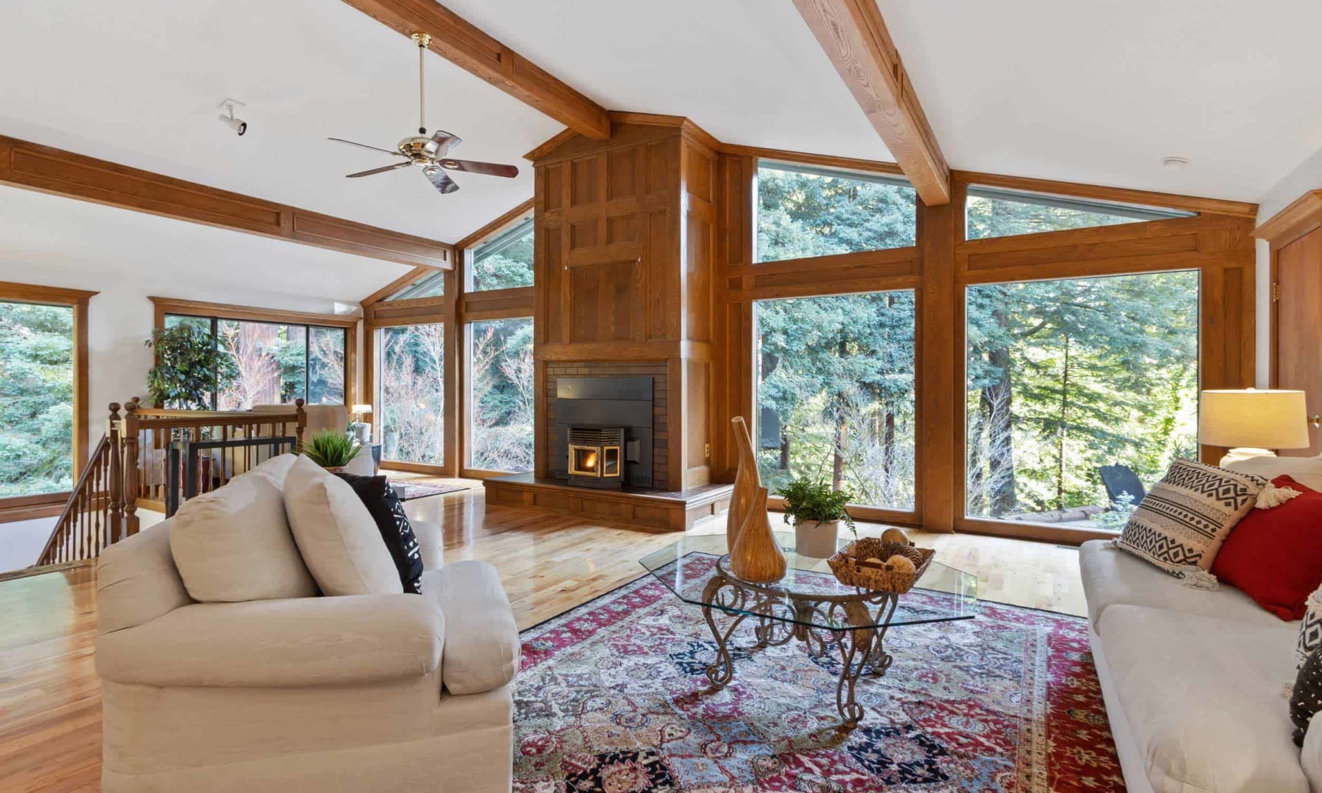 Living room with a timber-beamed ceiling, central fireplace, and tall windows overlooking trees.