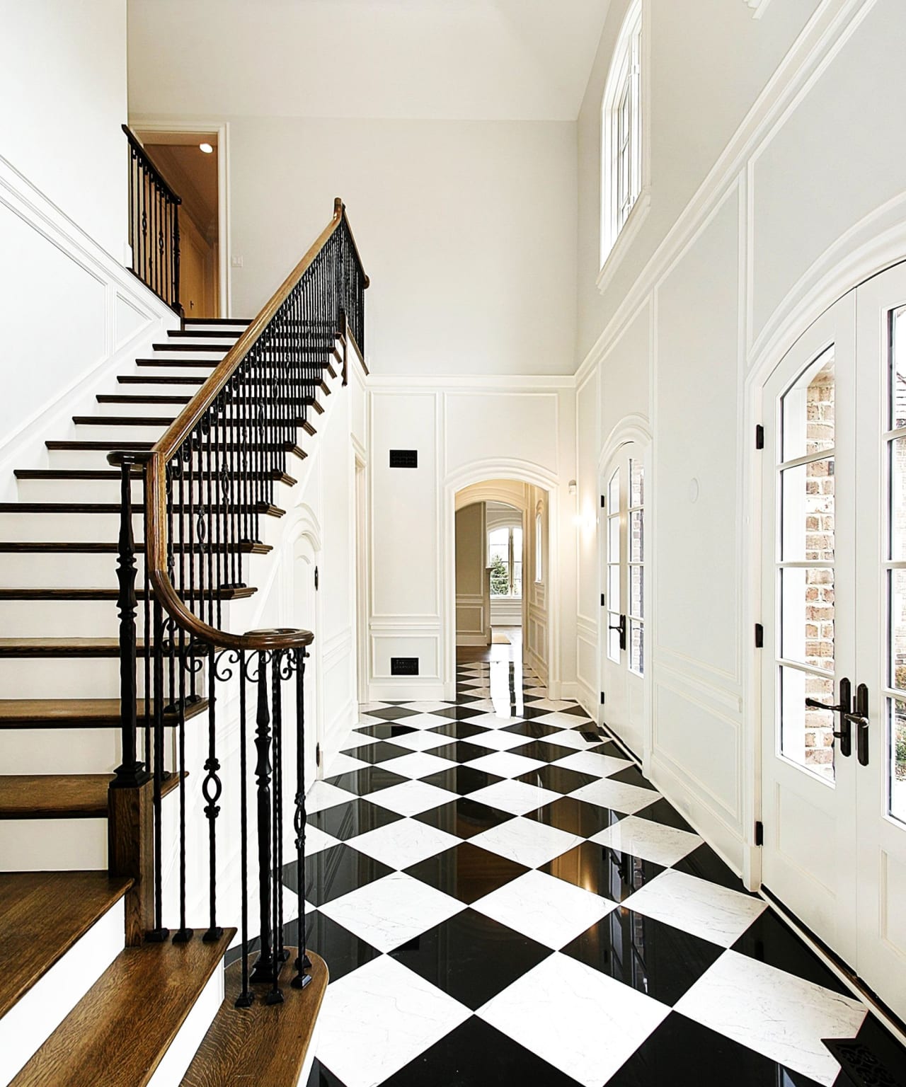 A black and white checkered floor stretches down a long hallway with beige walls and a wooden banister.