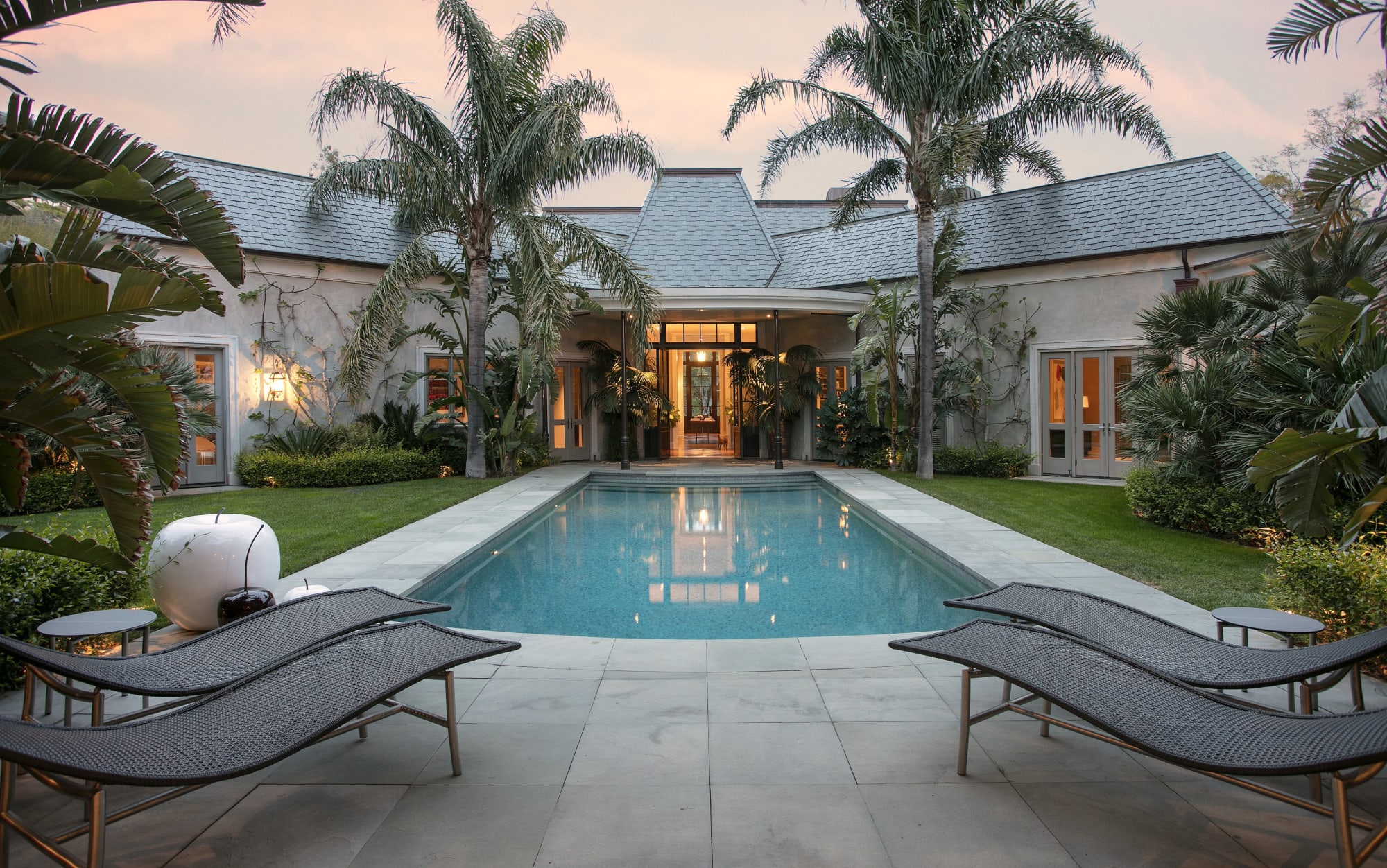 Home and swimming pool framed by palm trees and loungers at dusk.