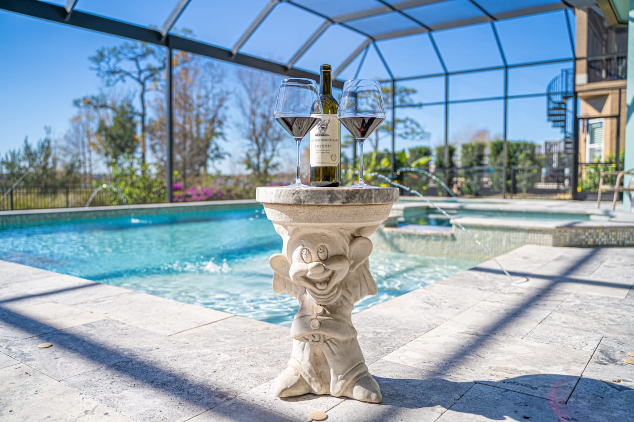 A wine bottle and two glasses sit on a table beside a pool, surrounded by lush greenery and a patio.