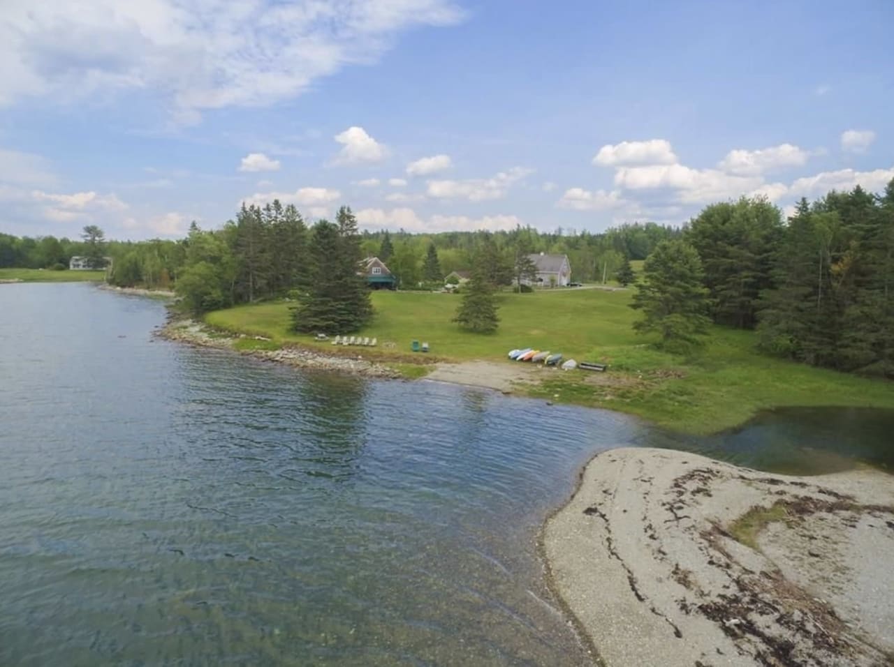 A lake with boats on the shore and a house, trees, and lush green grass in the background.