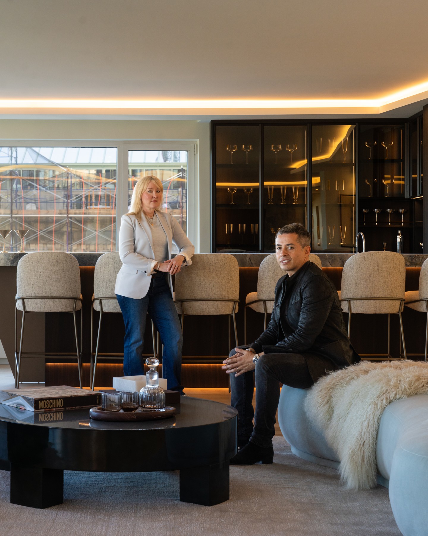 Mallory standing beside the bar as Brandon looks toward her from the sofa at 200 Central Park South