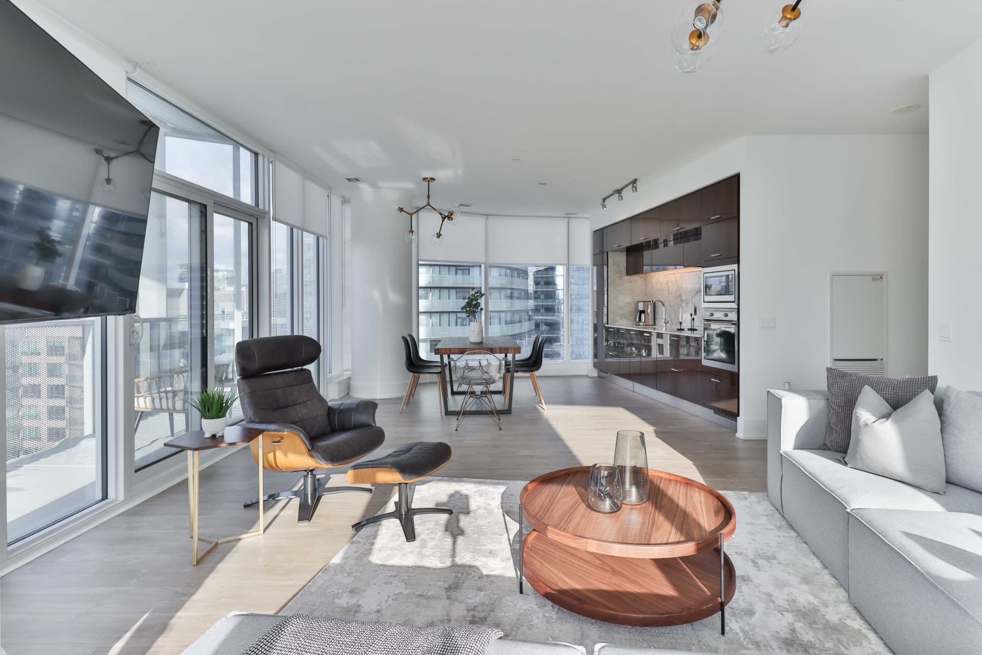 A modern open-plan living room with outside-view windows, flowing into a dining room and kitchen.