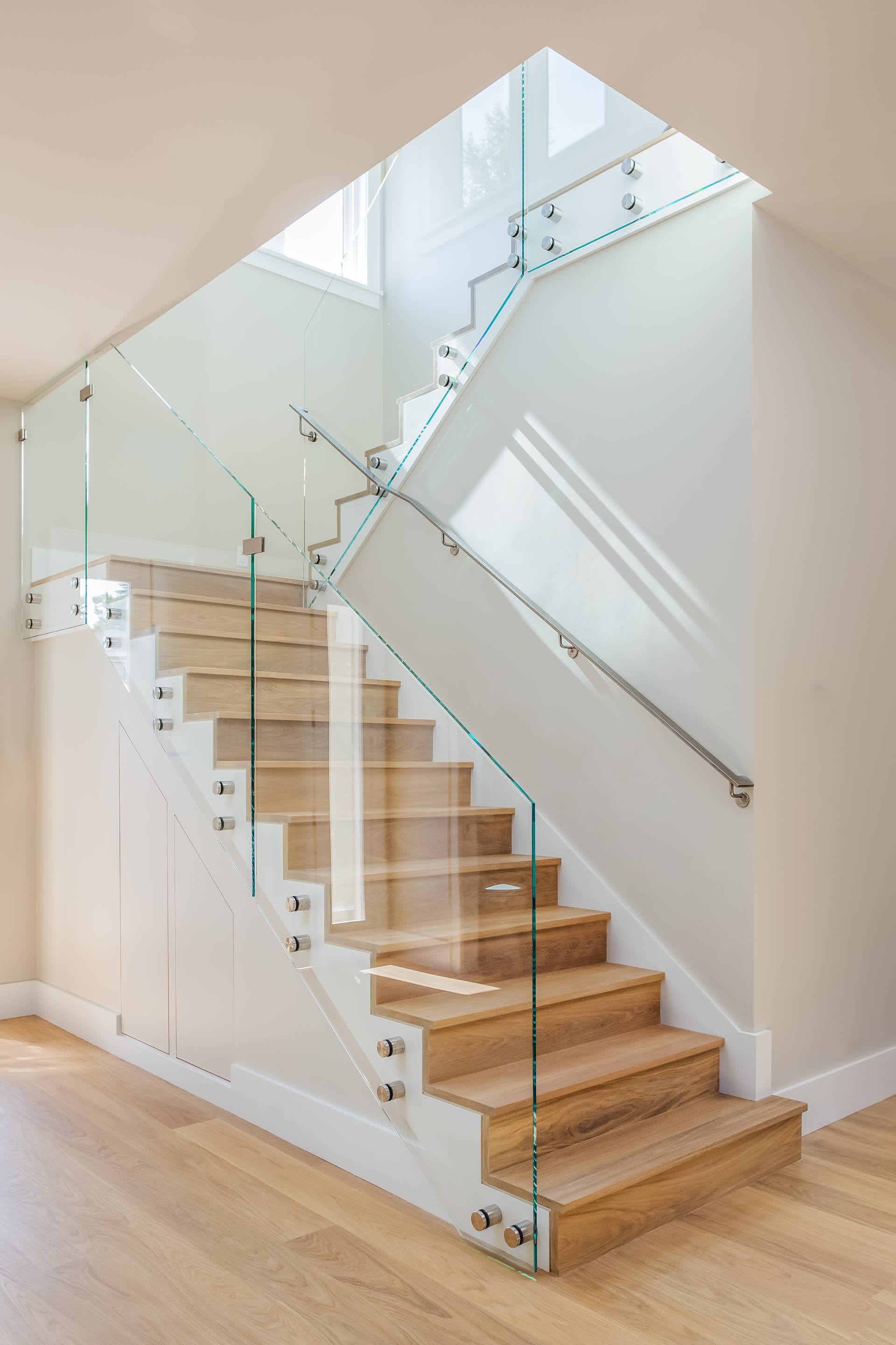 A close-up of a staircase with light-colored wooden steps and a modern glass railing system