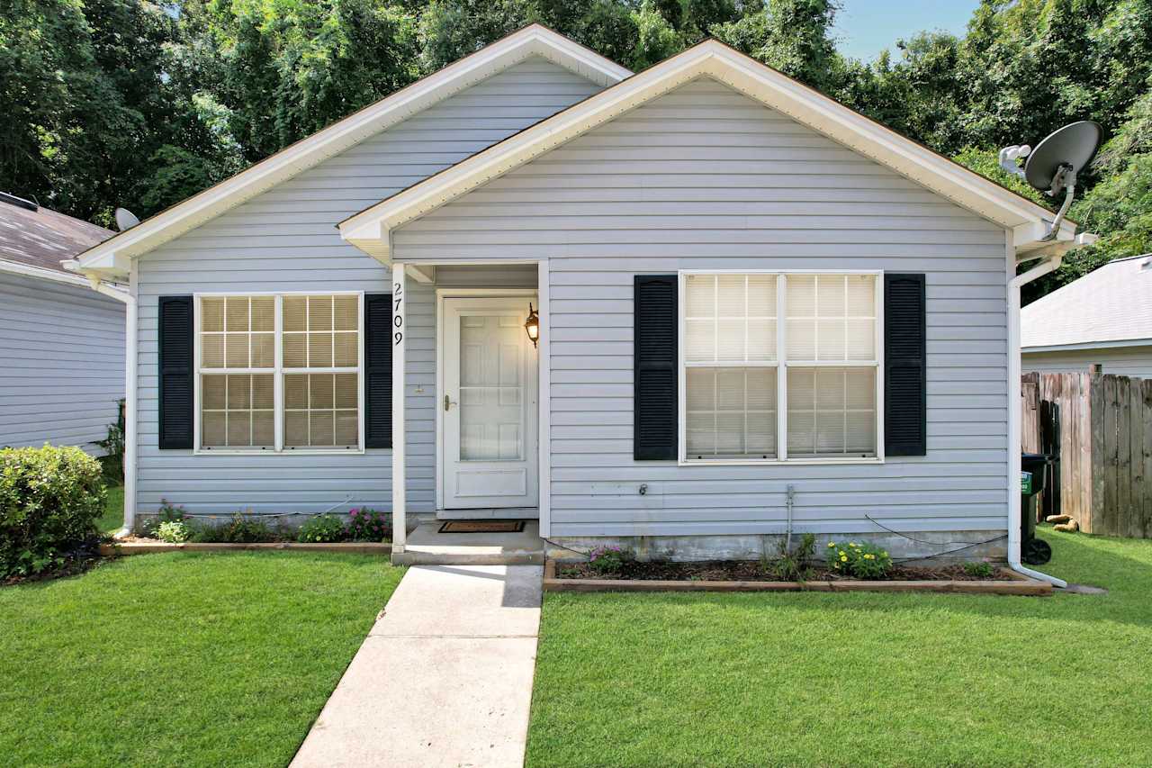 Front photo of home featuring lightly colored vinyl siding, white window grids, a white front door, black shutters, flowering garden beds, green grass, and a concrete walkway to the front door  at 2709 Oak Park Court, Tallahassee, Florida 32308