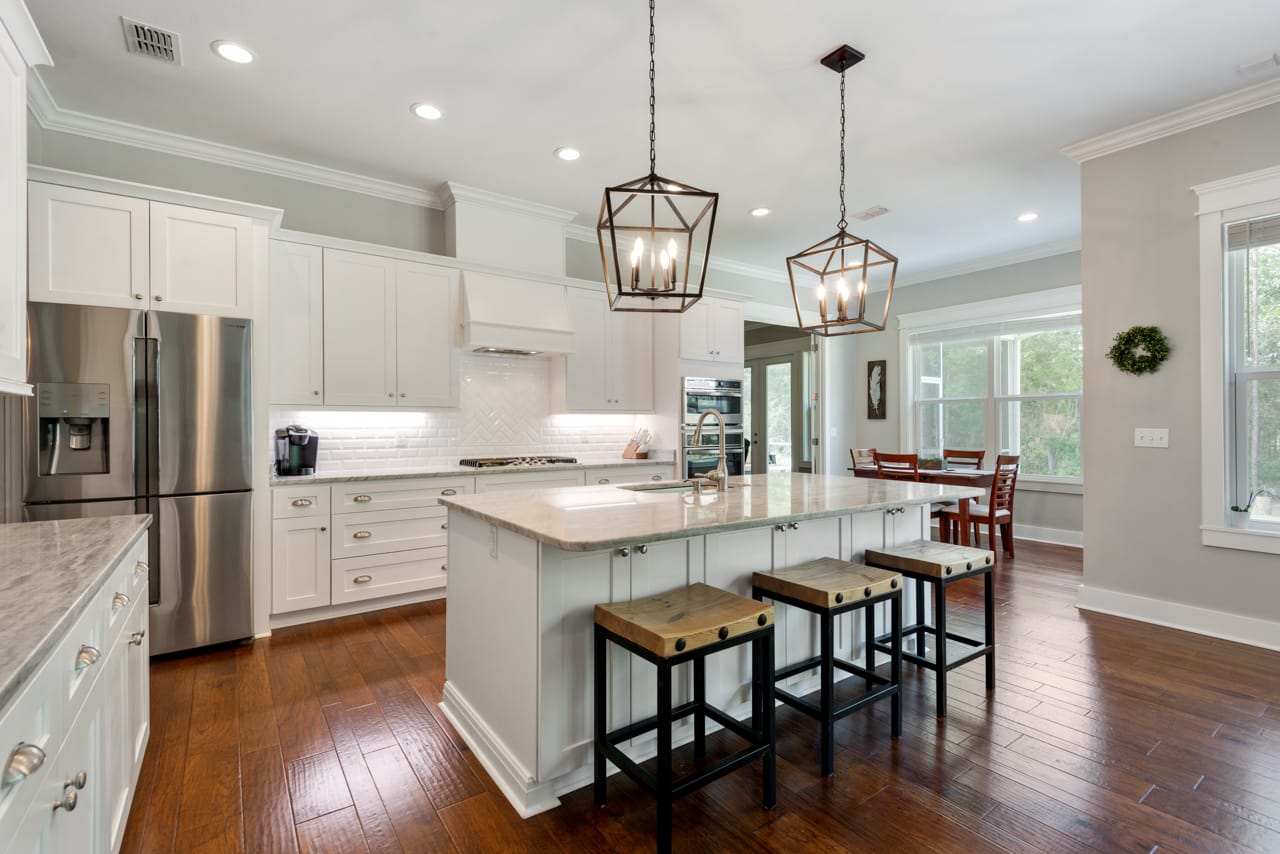 A modern kitchen with white cabinets, a large island with seating, and wooden flooring. The kitchen is bright and airy, with pendant lighting over the island.