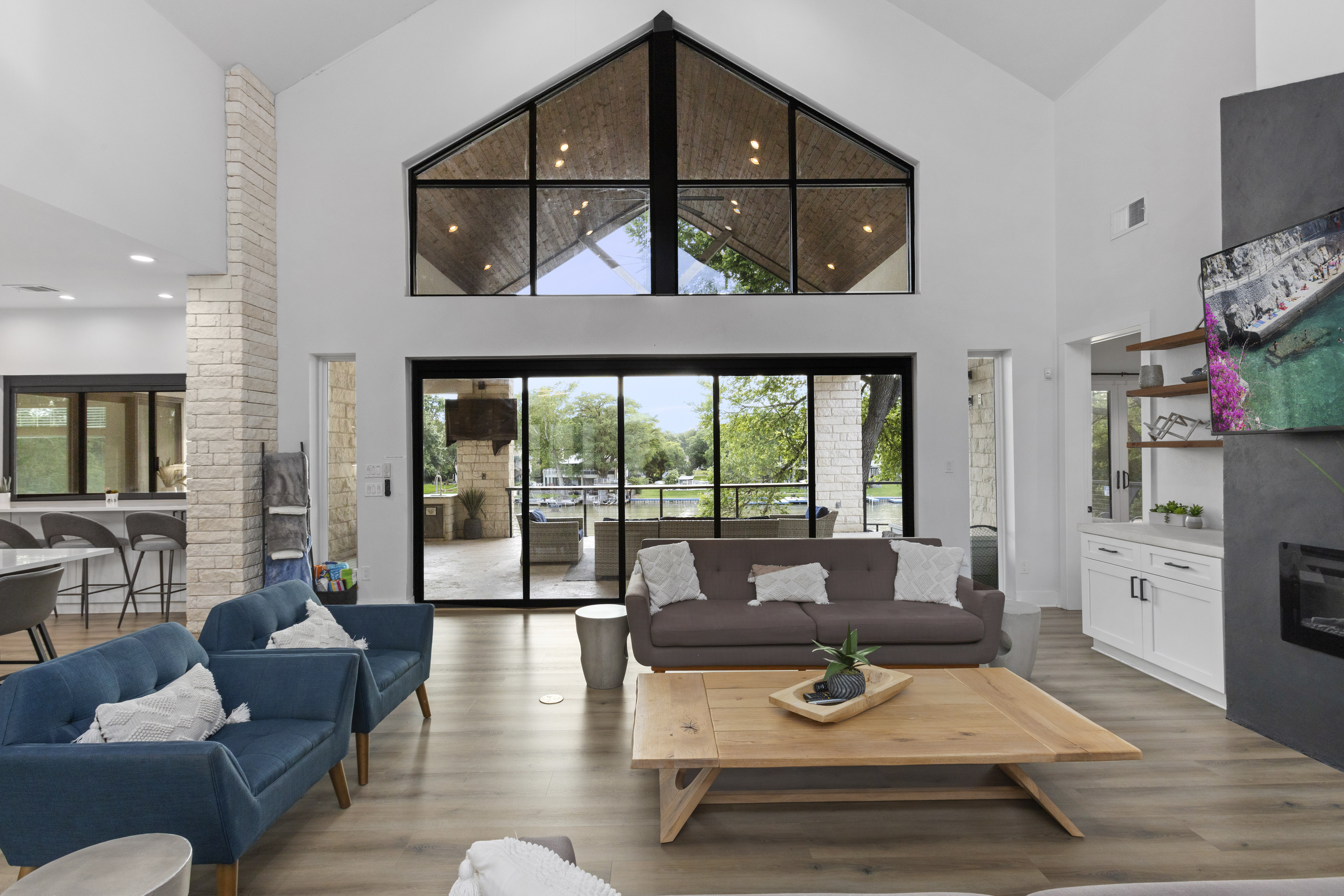 Great room with a vaulted white ceiling, a peaked window wall above black-framed sliding glass doors that open to a covered patio and the lake, a gray sofa, blue armchairs, a wood coffee table and a fireplace wall at right