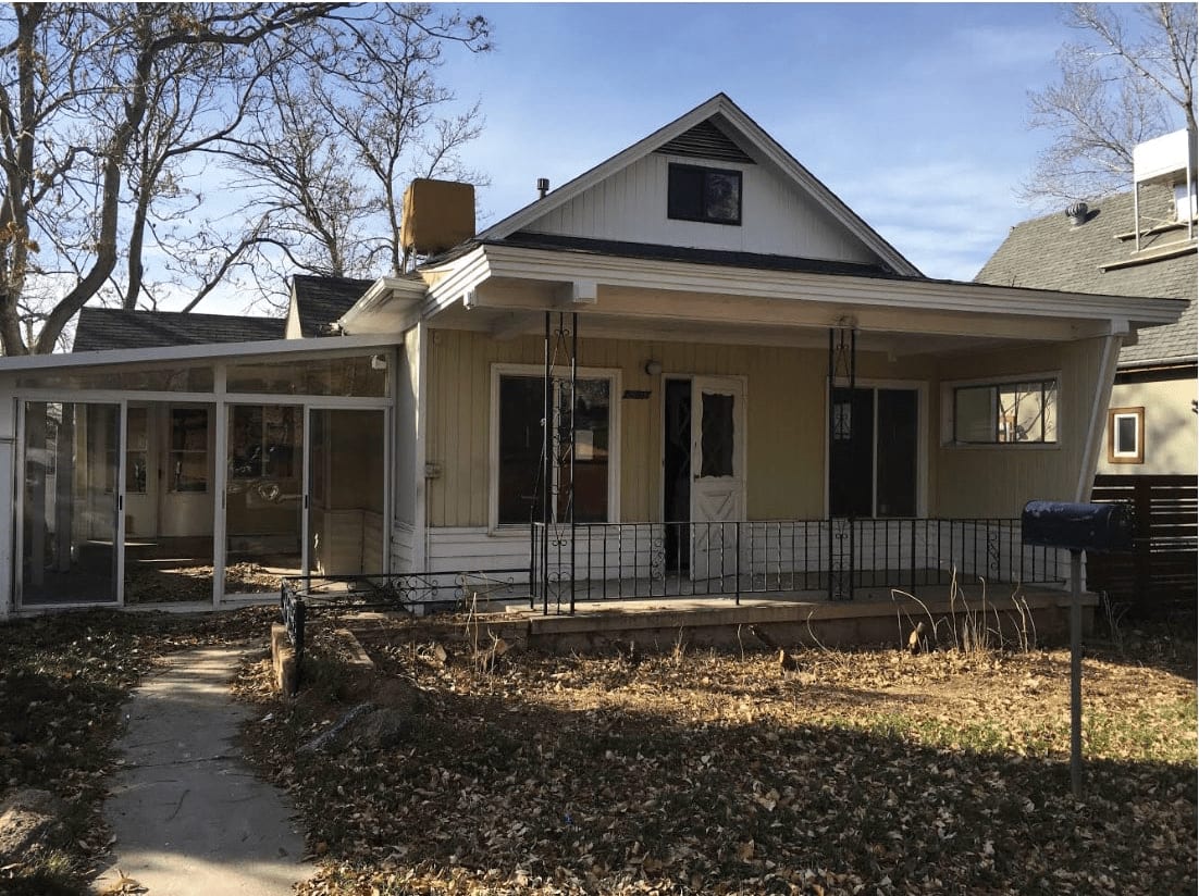  A quaint, older single-story house with light yellow siding and a white-trimmed gable roof. 
