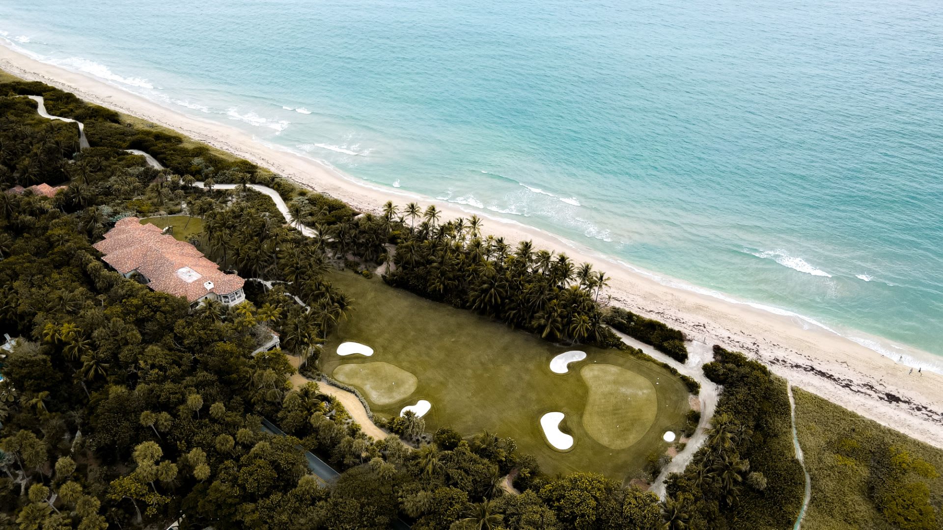 Aerial view of a beachfront estate with a palm-lined golf green beside turquoise water.