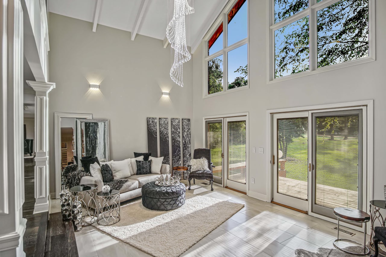 An interior view of a modern living room with high ceilings, large windows, and contemporary furnishings. The room features light-colored walls and floorings.