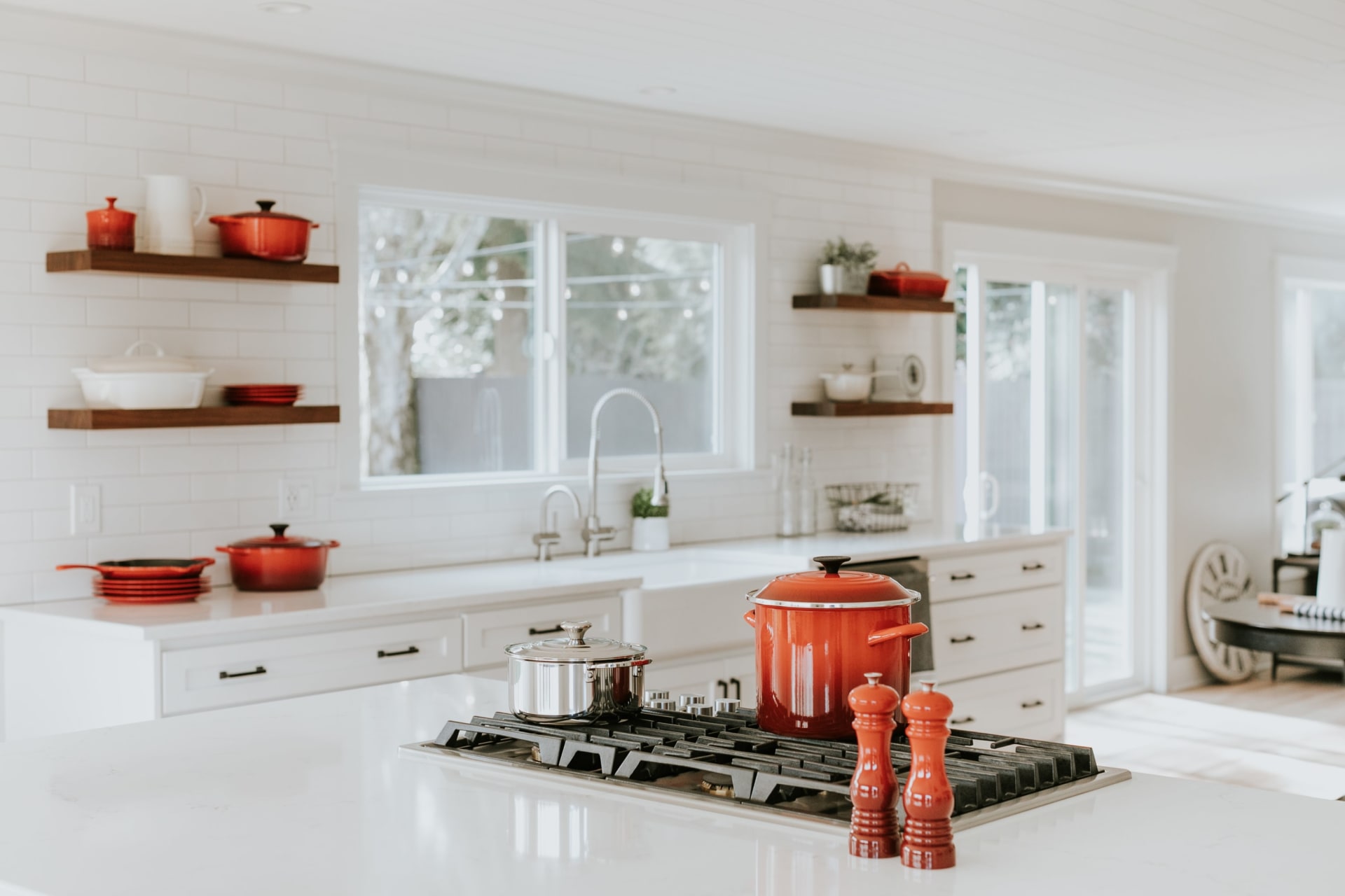 Bright white kitchen with open wood shelves, large windows, and red cookware on the island.
