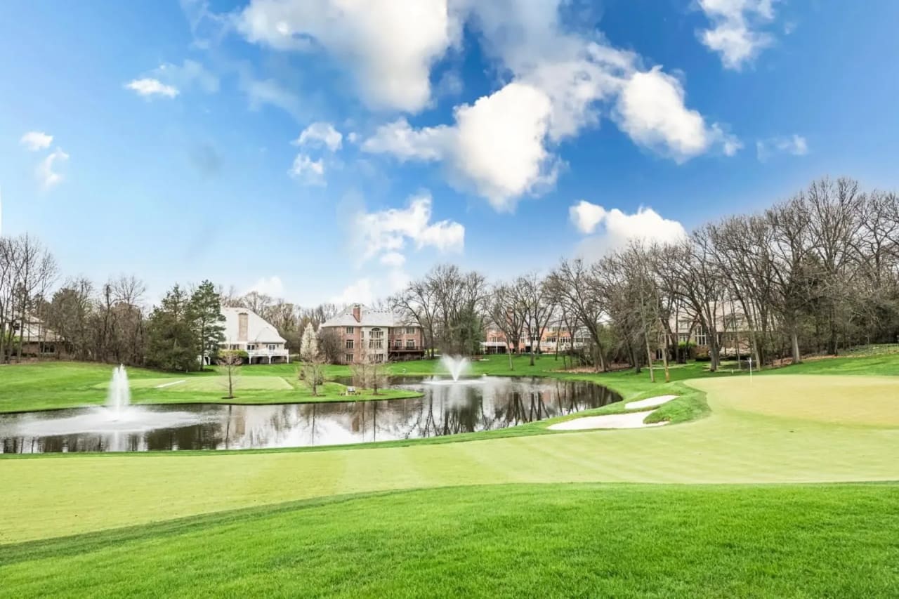 Scenic view of a golf course with a pond, fountains, and luxury homes in the background under a partly cloudy sky.