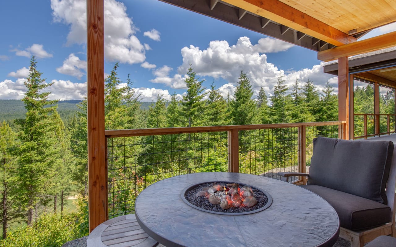 A covered outdoor patio with a round fire pit and a chair, overlooking a scenic view of a forest and mountains