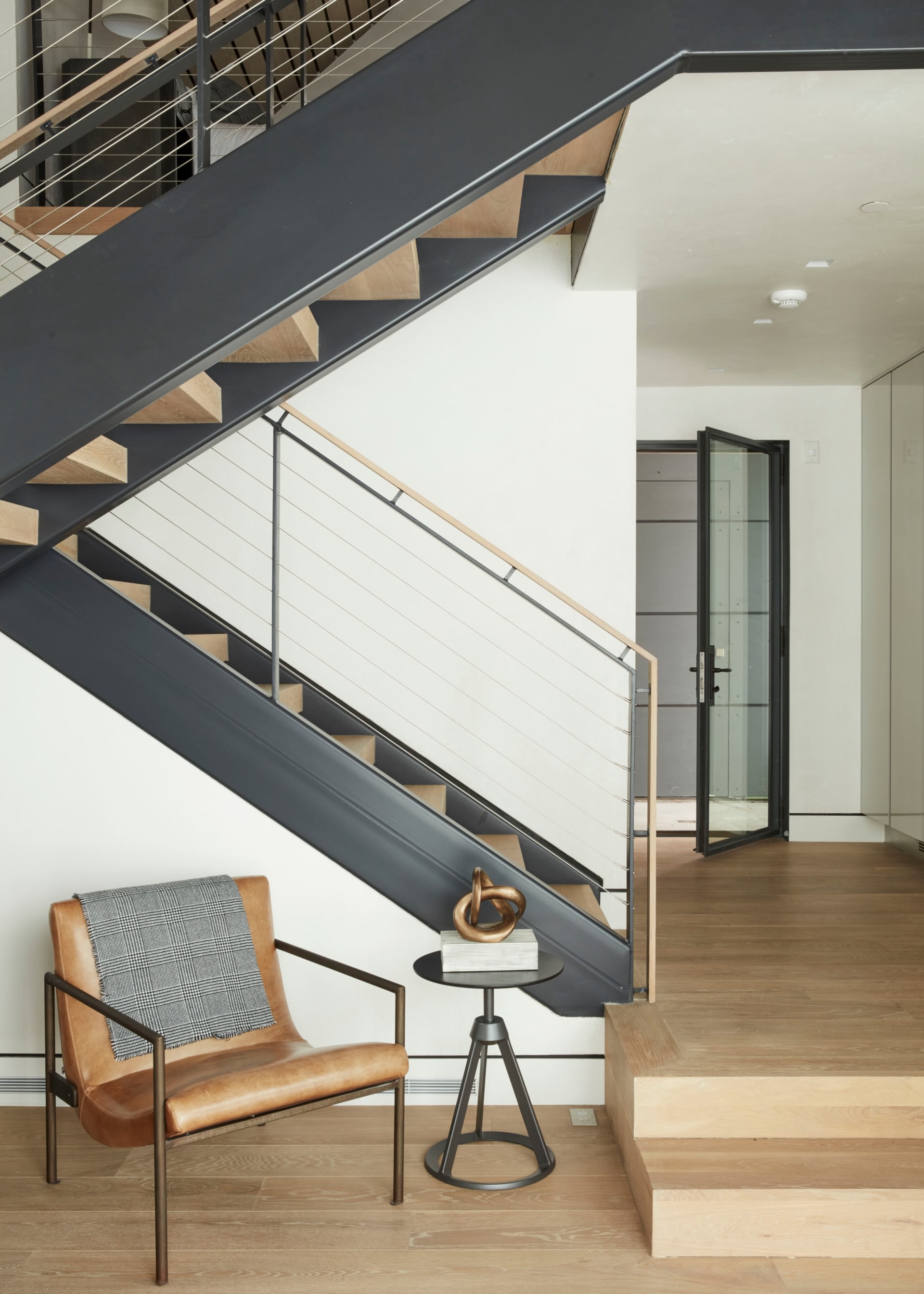 Underneath the stairs, there's a light brown leather and metal armchair with a patterned throw blanket