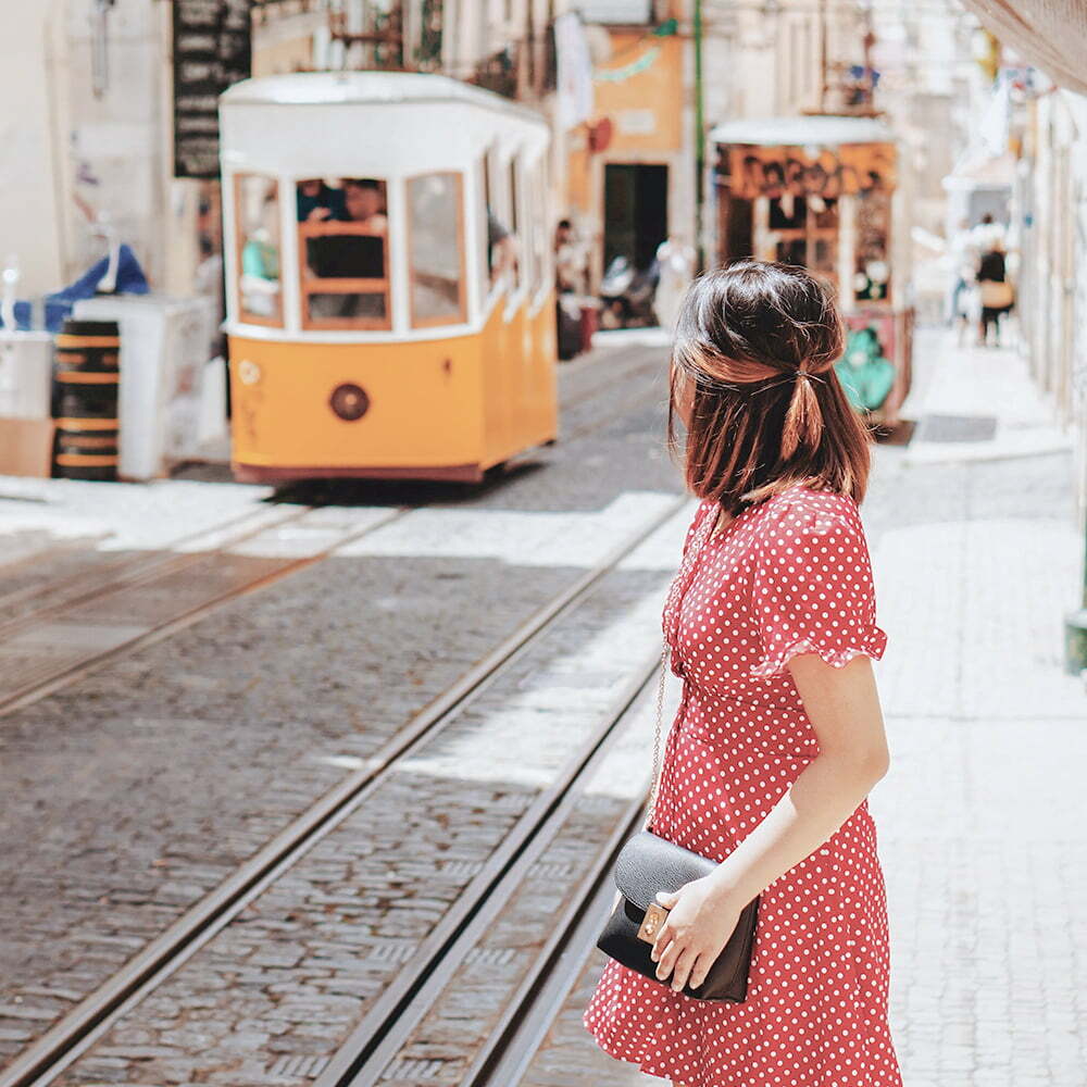 Woman in a red polka-dot dress watches a yellow tram on a cobbled street.