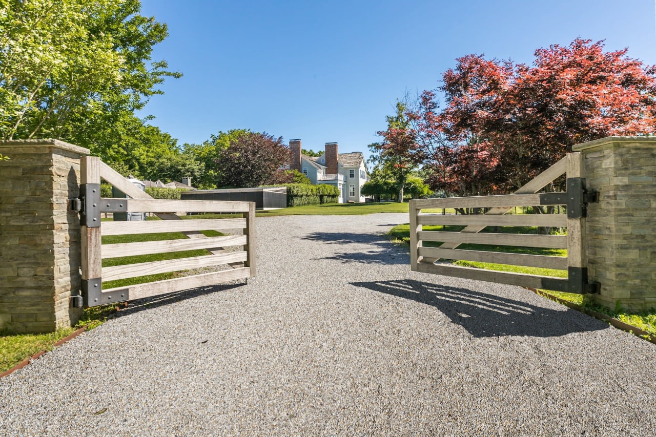 Double wooden gate at dawn or dusk, opening to a tree-lined driveway with two large, traditional houses in view.