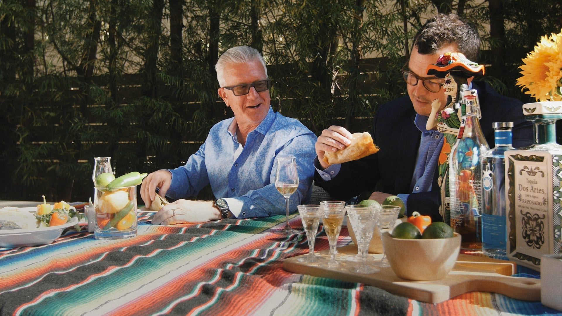  Two men are seated at an outdoor table covered with a colorful striped blanket.