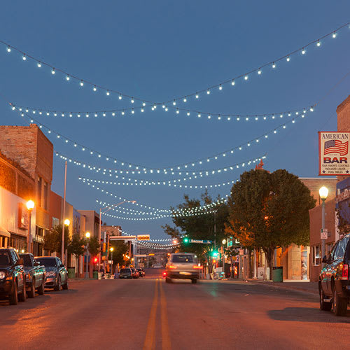 A small town street at night with parked cars and lit-up buildings.