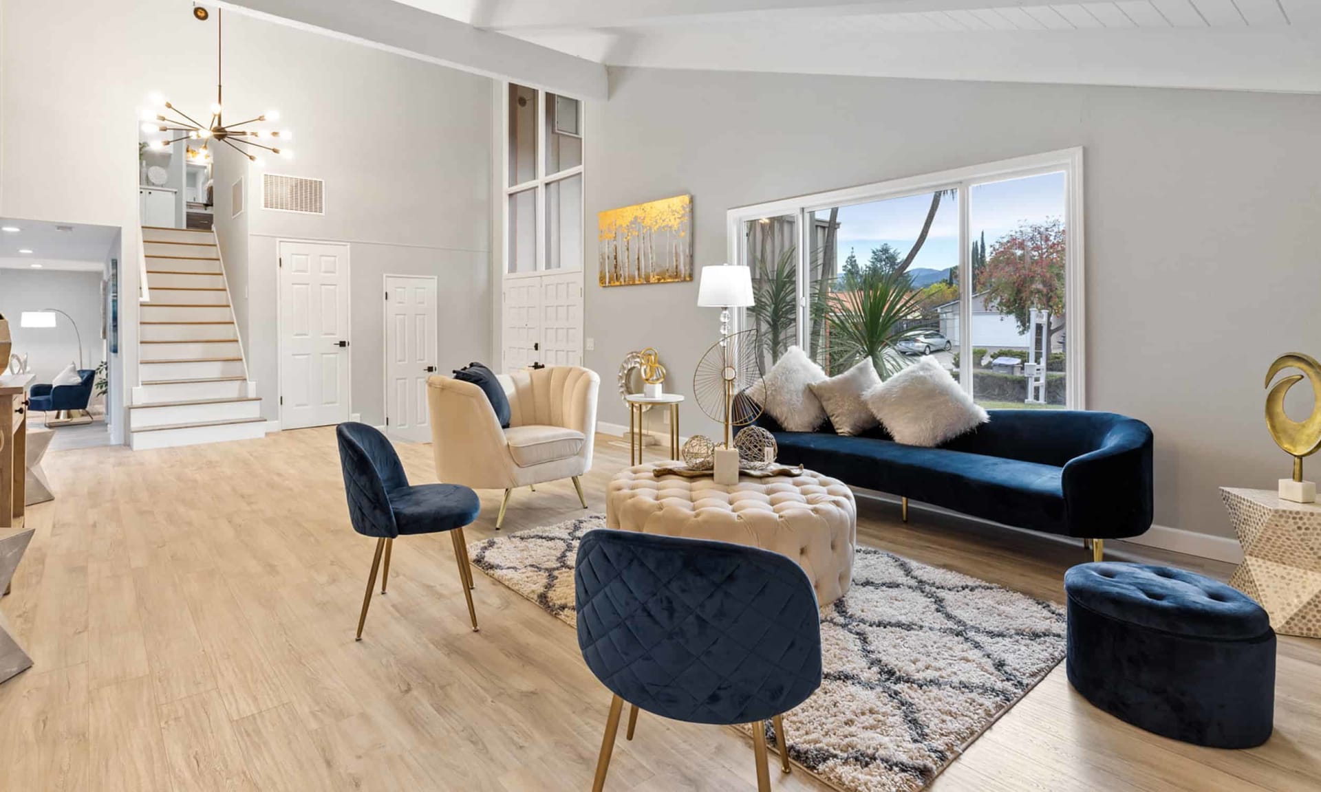 Furnished living room with a vaulted ceiling, staircase, and blue sofa beside large windows.