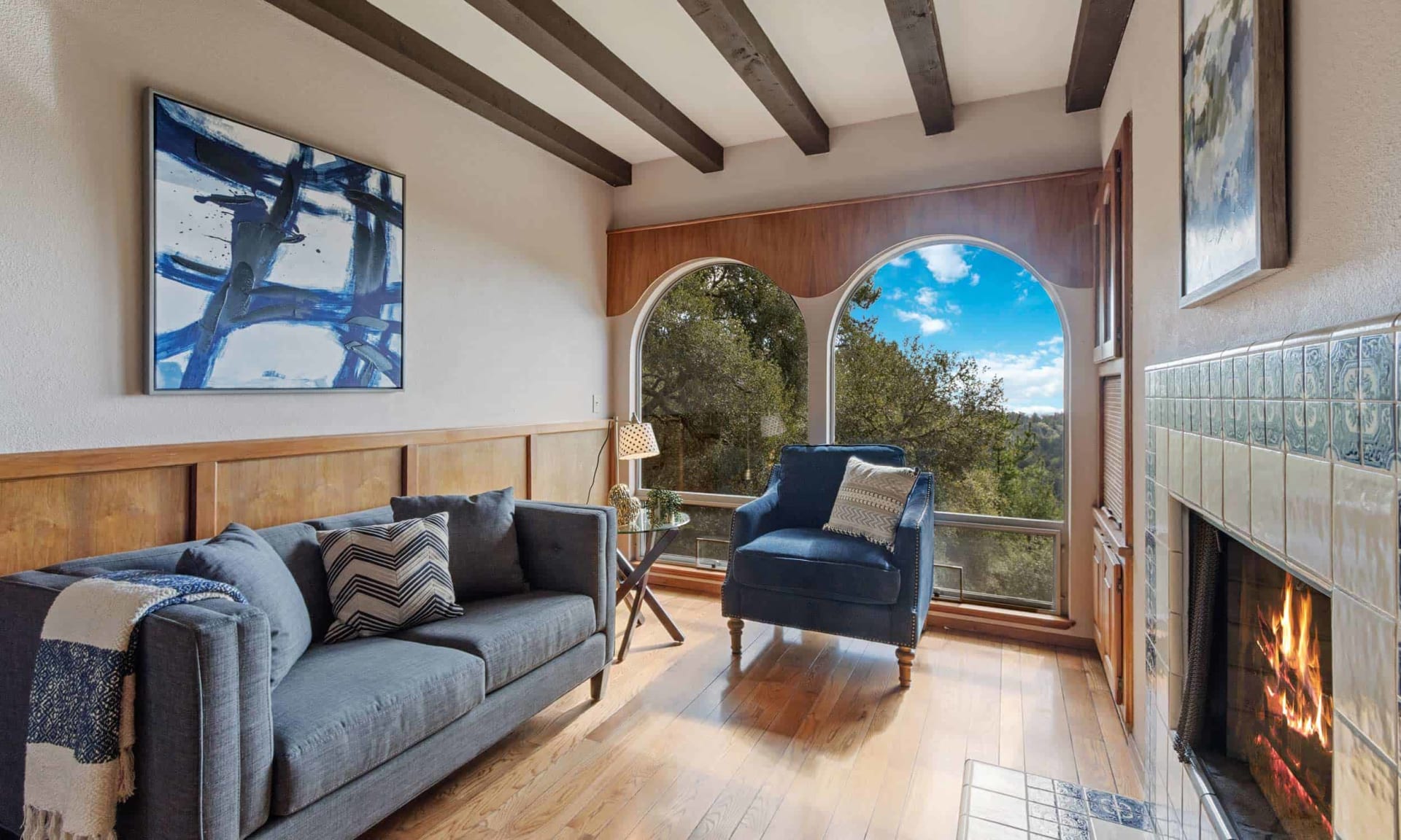 Living room with arched windows overlooking trees, blue seating, exposed beams, and a tiled fireplace.