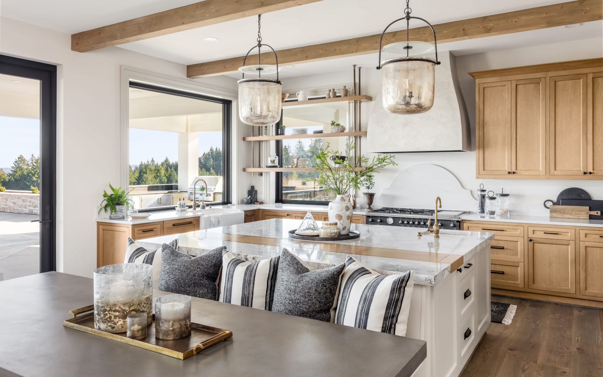 Bright kitchen with a marble island, wood cabinetry, exposed beams, and windows overlooking trees.