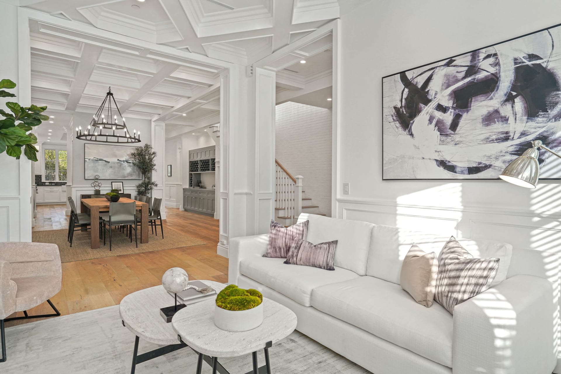  A brightly lit, expansive open-concept living space features a white sofa with throw pillows in the foreground, facing two round marble-top coffee tables. 