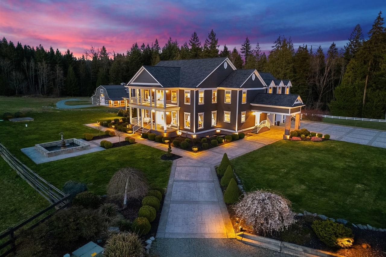 A large, two-story house with a wraparound porch, illuminated by warm lights against a dramatic twilight sky